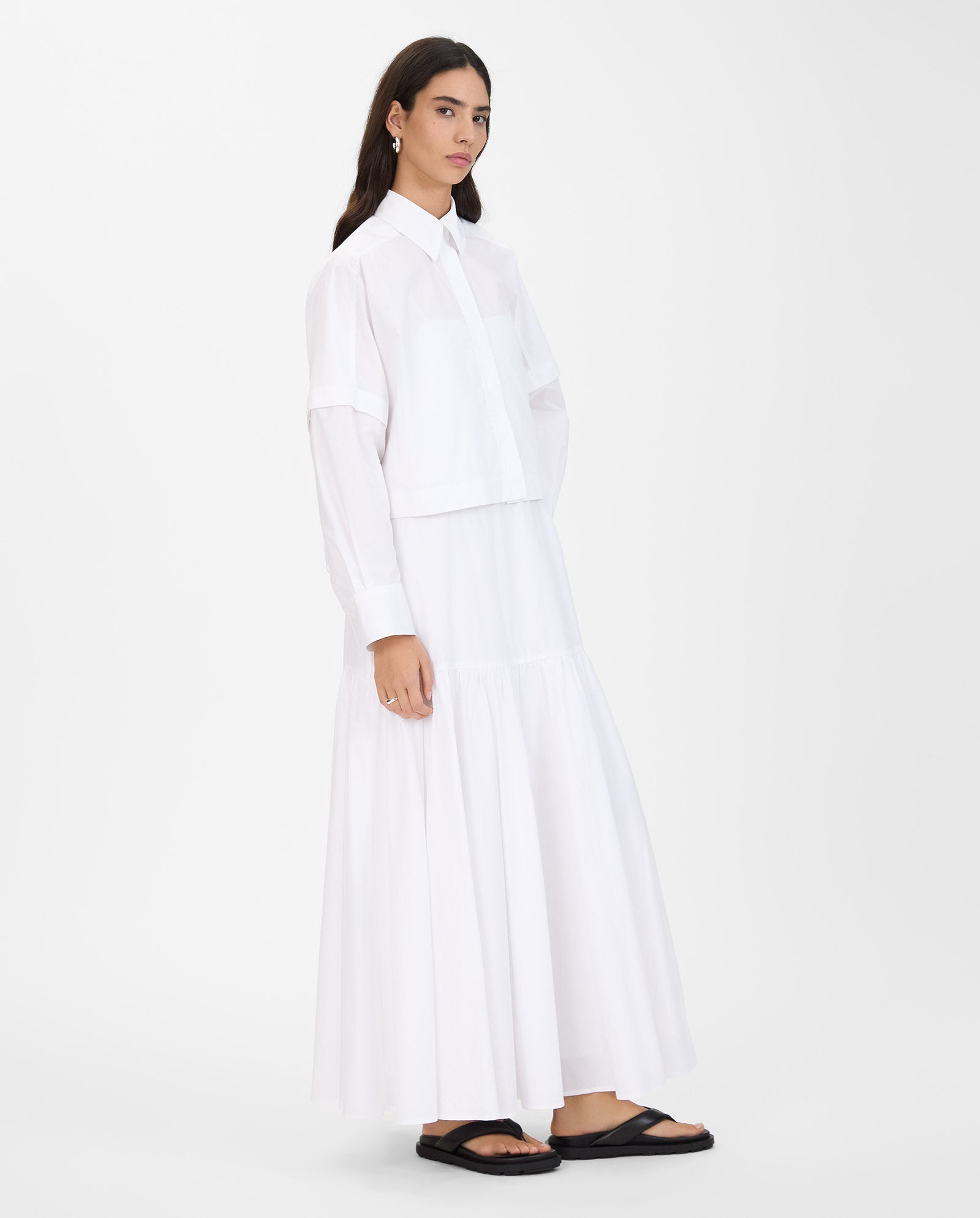 A woman wears a long white dress, the IVY OAK ELVIRA Shirt with detachable sleeves, and black sandals, standing against a plain white background.