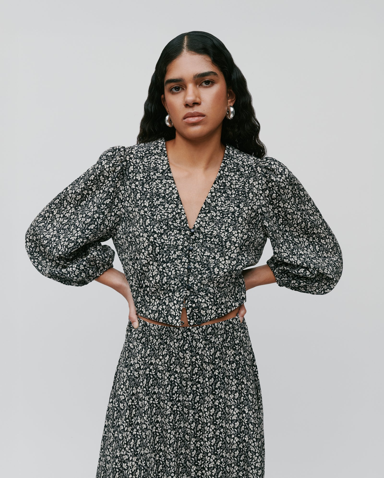 A woman in the IVY OAK ENRICA Blouse, featuring a black floral design with a V-neck and 7/8 balloon sleeves, stands with hands on hips against a plain background.