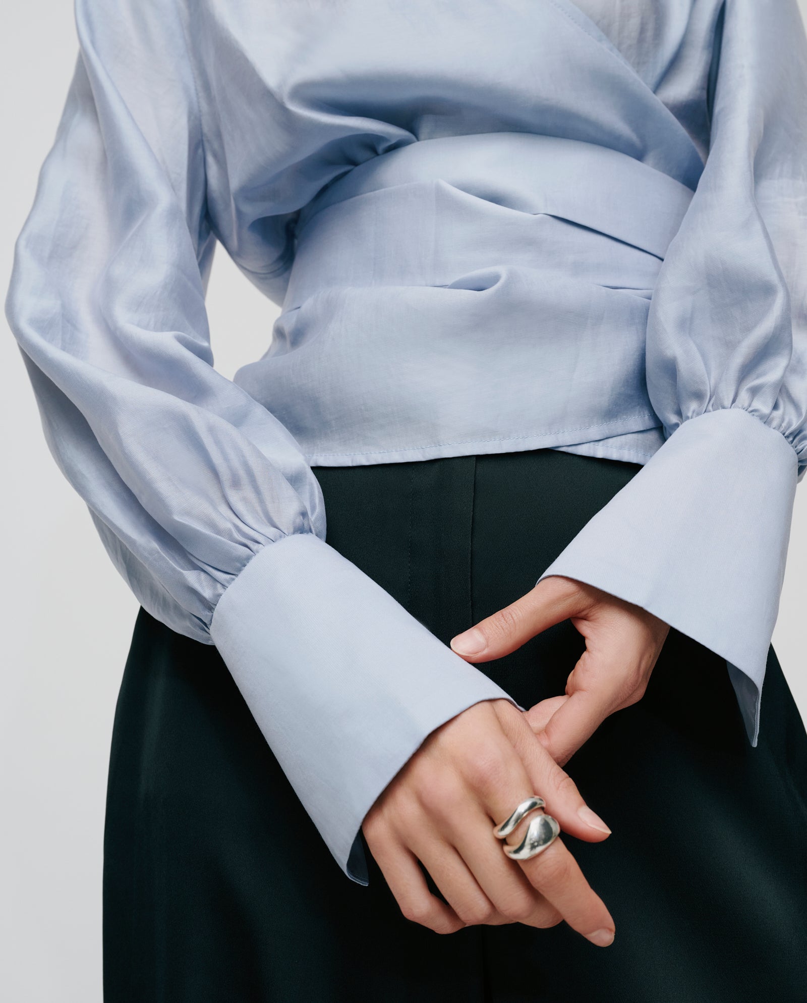 A person wears the IVY OAK ELOISE MAY Blouse in light blue organza with wide cuffs, paired with black pants and a chunky silver ring on one hand.