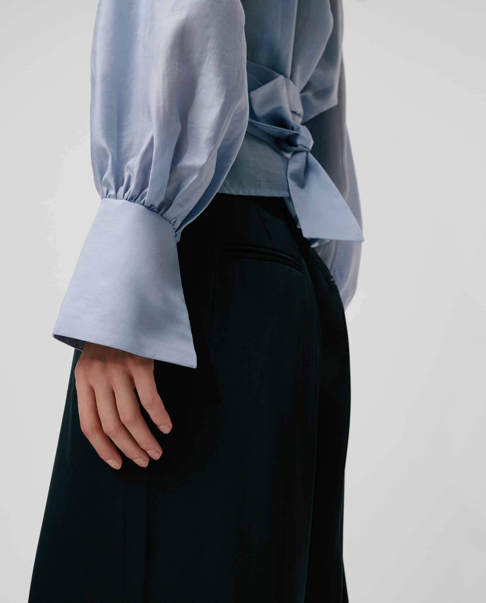 A person shown from the side wears the ELOISE MAY Blouse by IVY OAK, a light blue wrap top with flared cuffs and a long belt, paired with black pants.