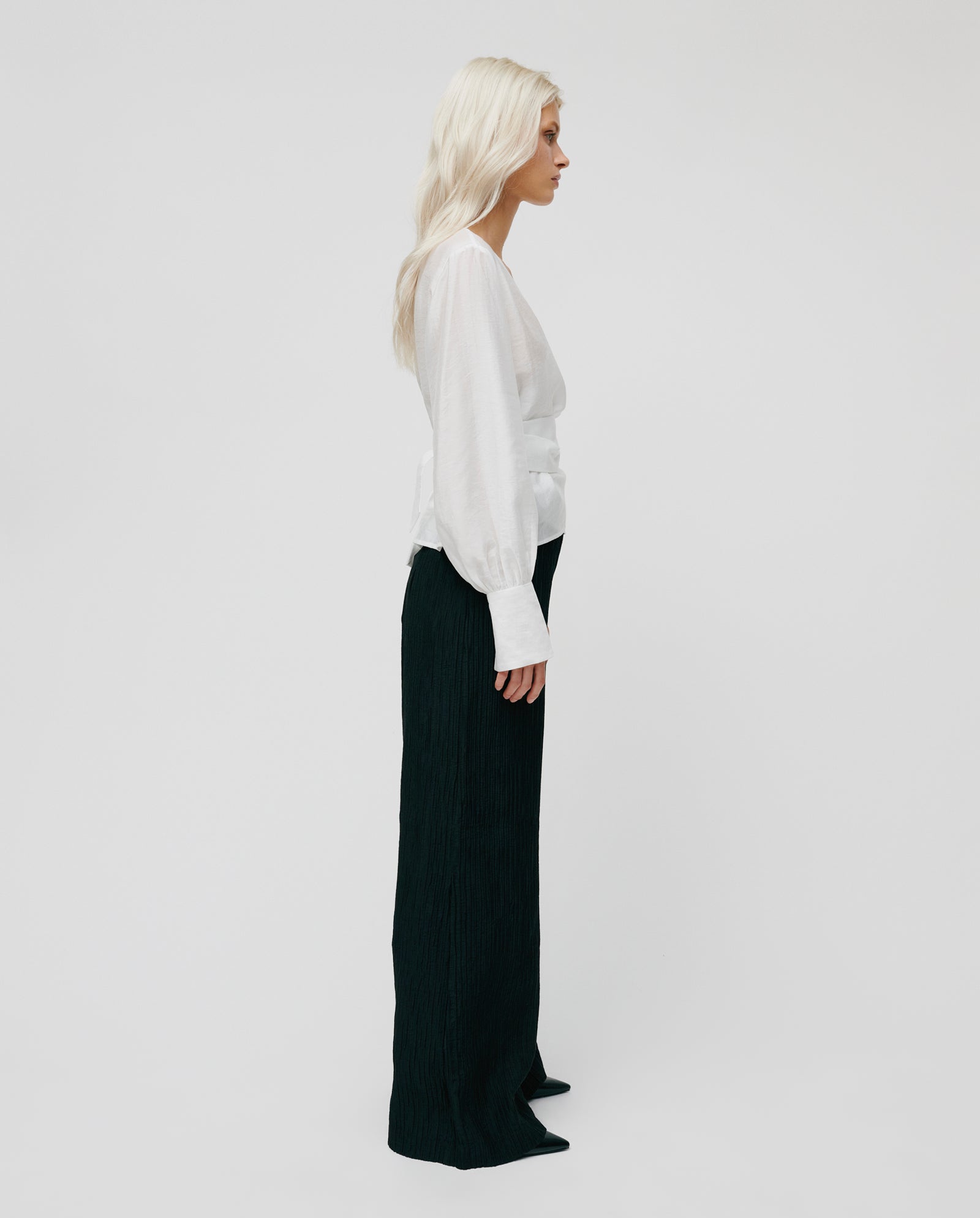 Woman with long blonde hair wears the IVY OAK ELOISE MAY Blouse—a white wrap blouse with long cuffs—paired with black wide-leg pants, standing in profile against a plain background.