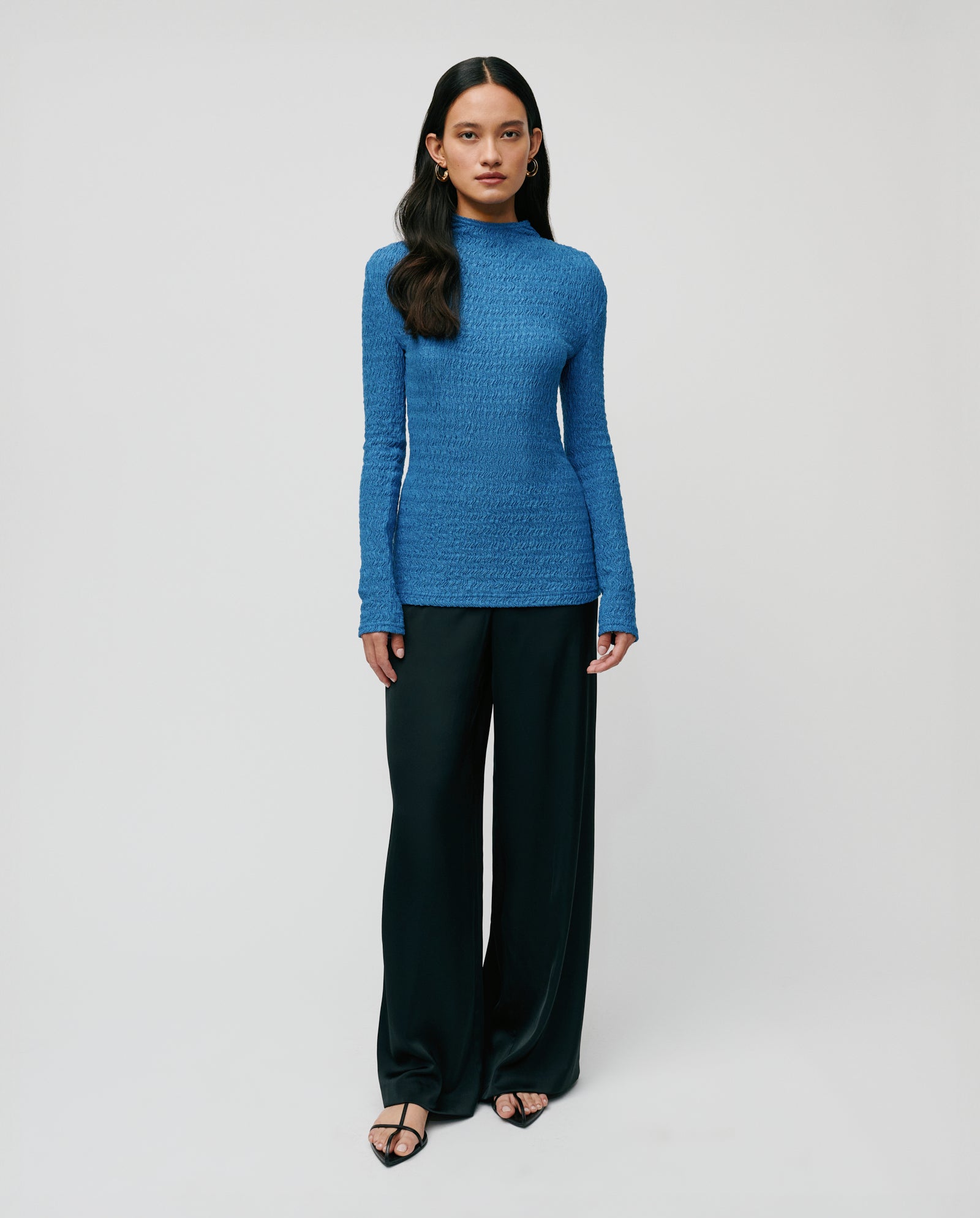 Woman wearing the IVY OAK TOSKA Top in blue, featuring long sleeves and a body-hugging fit, styled with black wide-leg pants against a plain background.