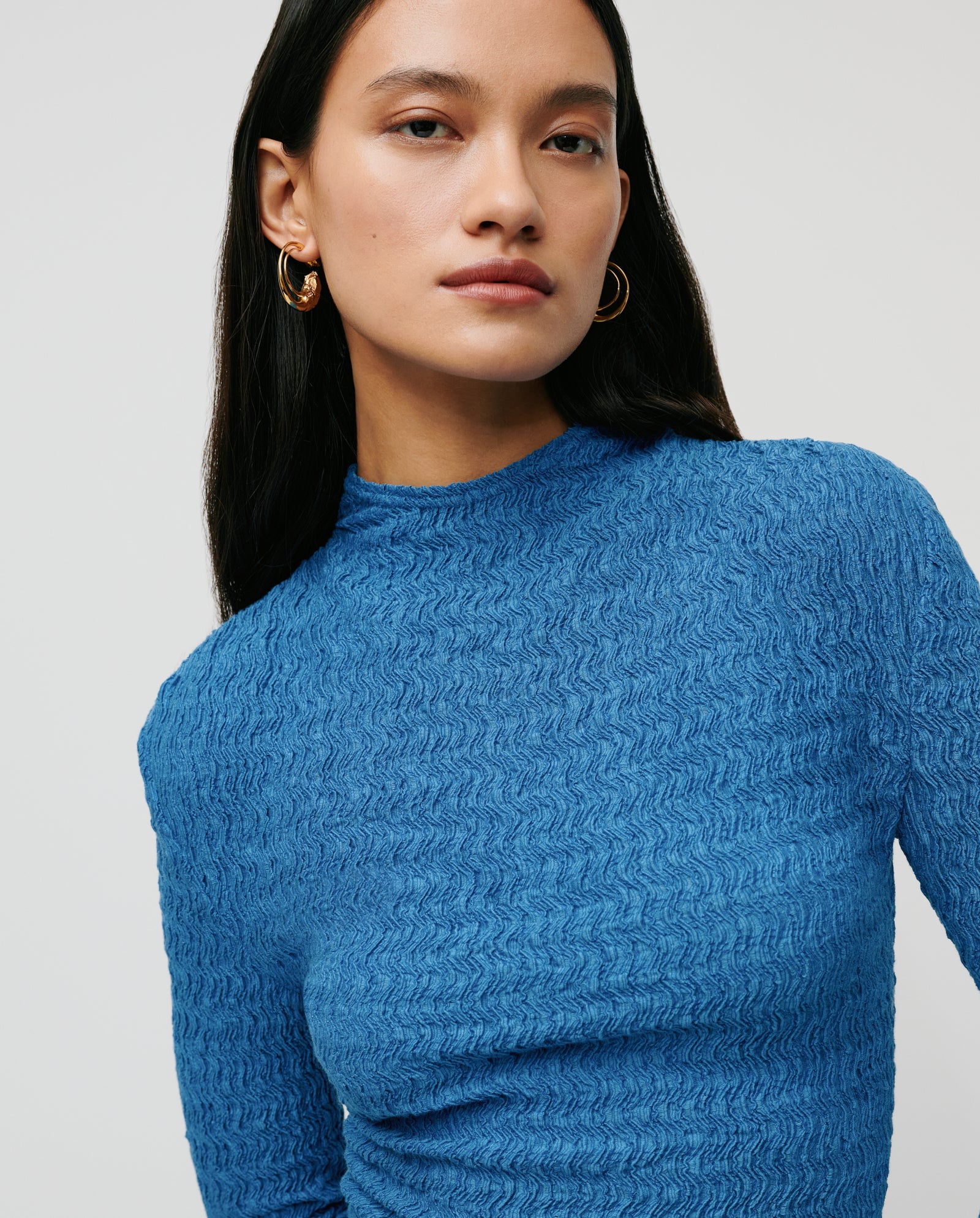A woman with long dark hair wears the IVY OAK TOSKA Top in textured blue with long sleeves and gold hoop earrings, confidently facing the camera.