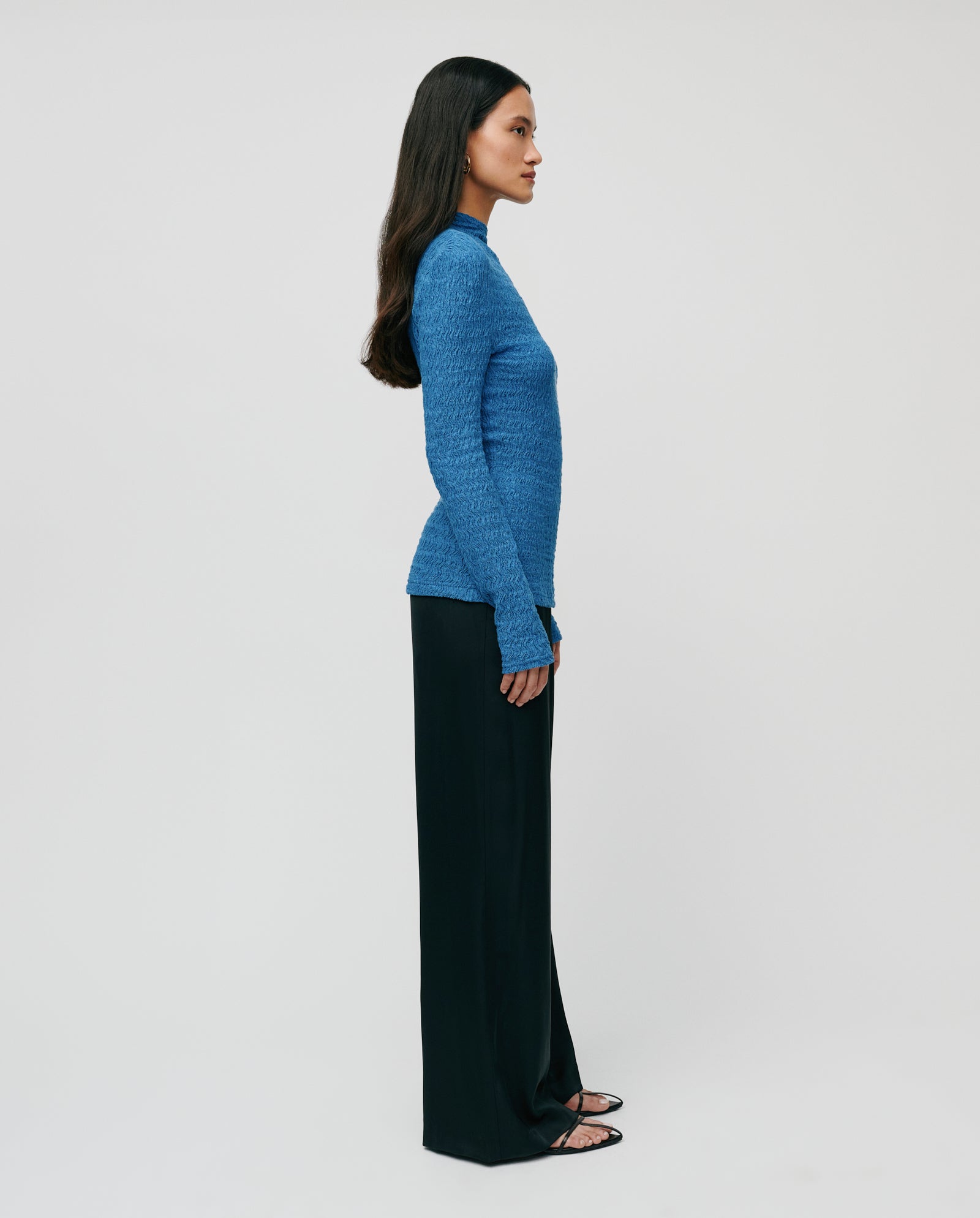 A woman stands sideways against a plain background, wearing a body-hugging IVY OAK TOSKA Top with long sleeves paired with black wide-leg pants.