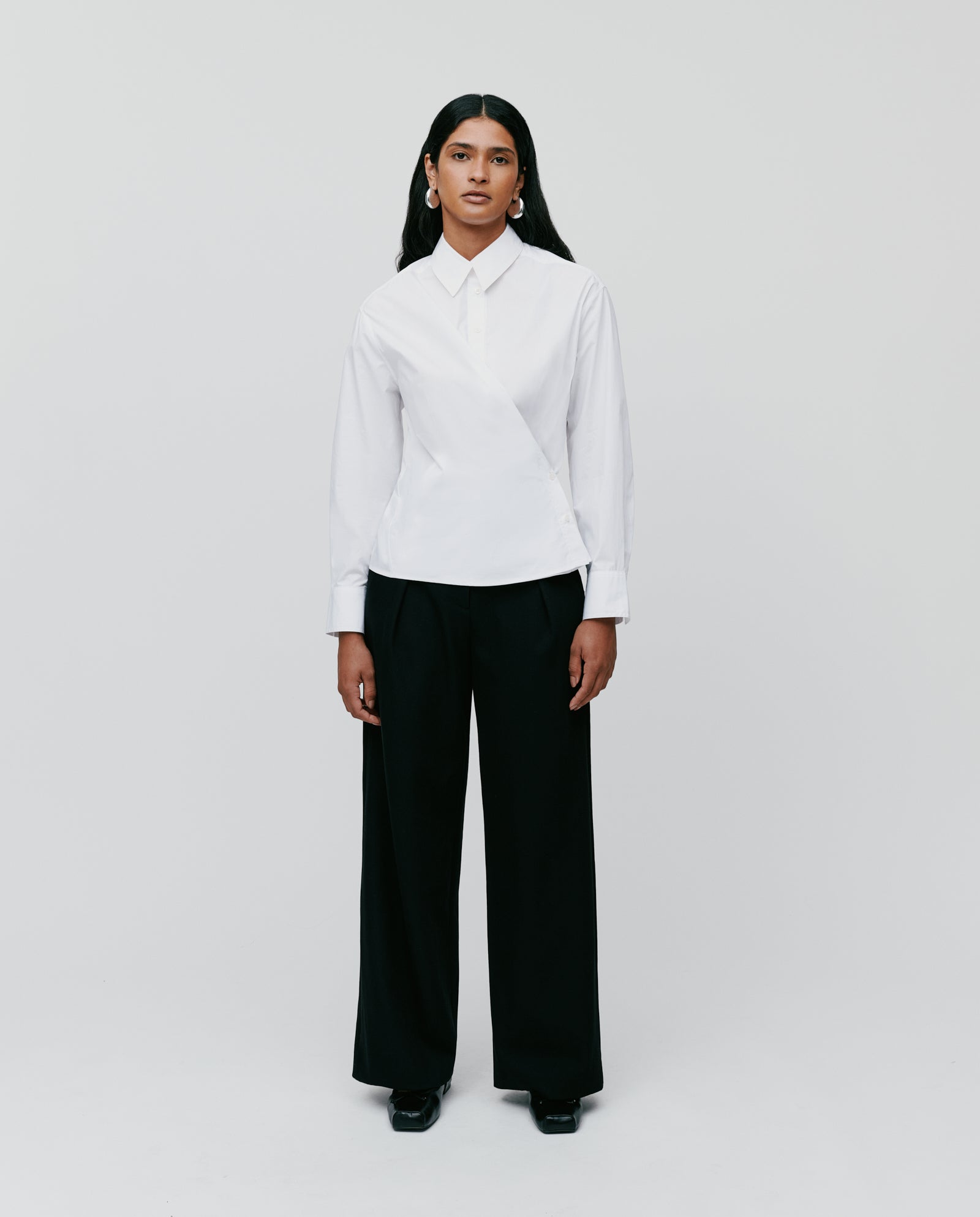 A person stands against a white background, wearing the oversized IVY OAK EVONNE Blouse with a button placket and loose black trousers.
