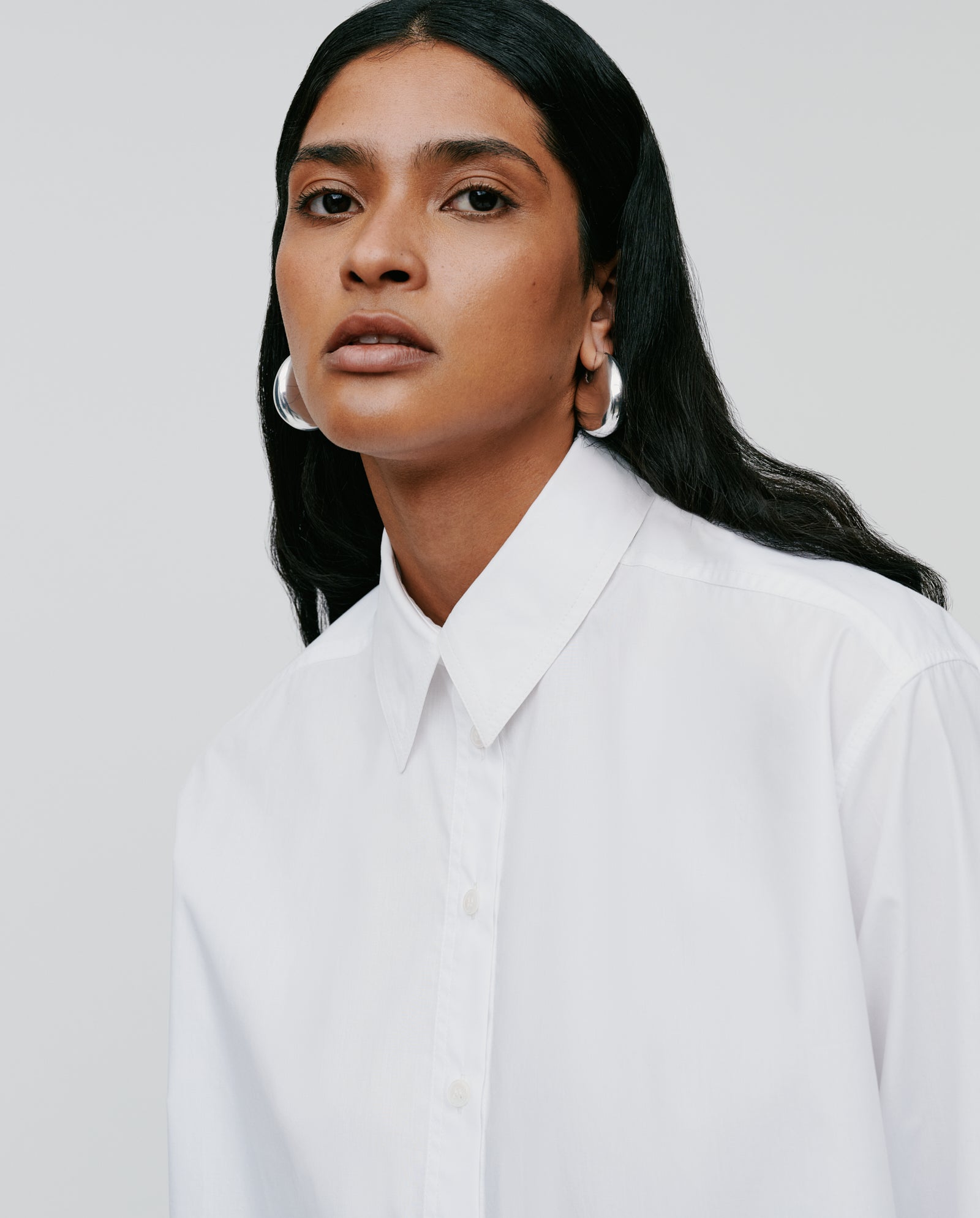 A woman with long dark hair wears the IVY OAK EVONNE Blouse, an oversized white top with a button placket, paired with silver hoop earrings against a plain background.