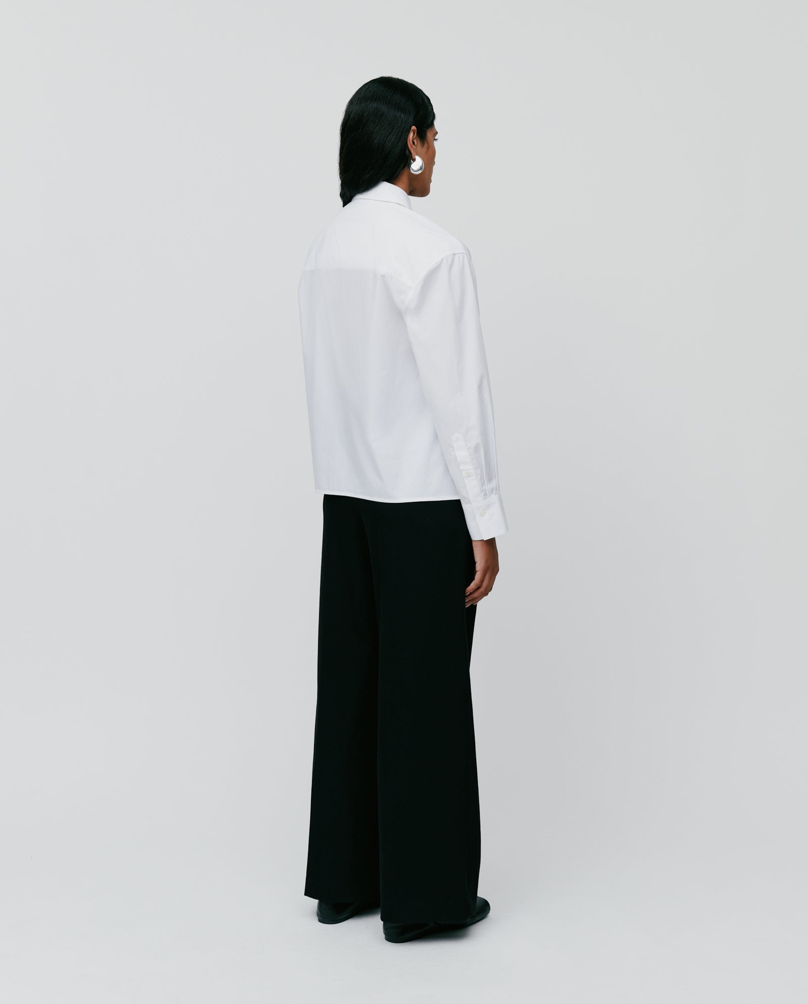 Person stands with back to camera wearing the IVY OAK EVONNE Blouse, featuring an oversized fit and button placket, paired with black wide-leg pants against a plain background.