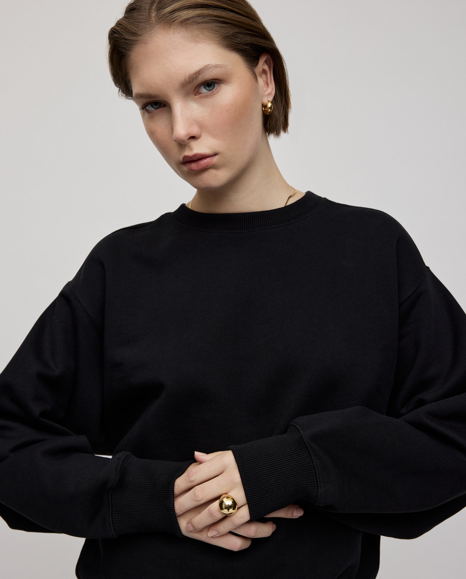 A woman with short hair wears an oversized black IVY OAK KAREN ROSE Jumper, paired with gold earrings and a ring, looking at the camera. For a closer fit, select a smaller size.