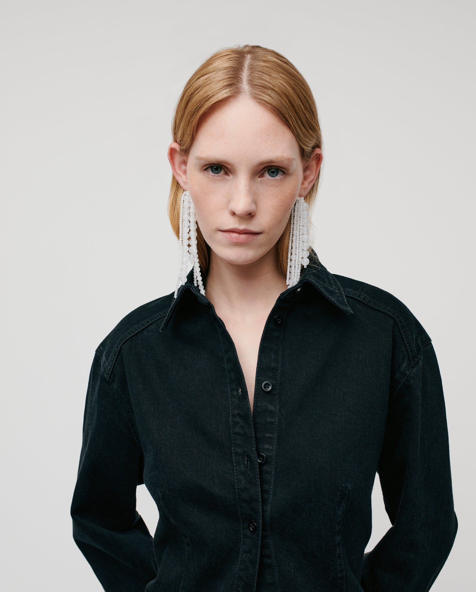 A woman with straight blonde hair wears the IVY OAK BERENICE Blouse in washed denim and large white statement earrings, looking at the camera.