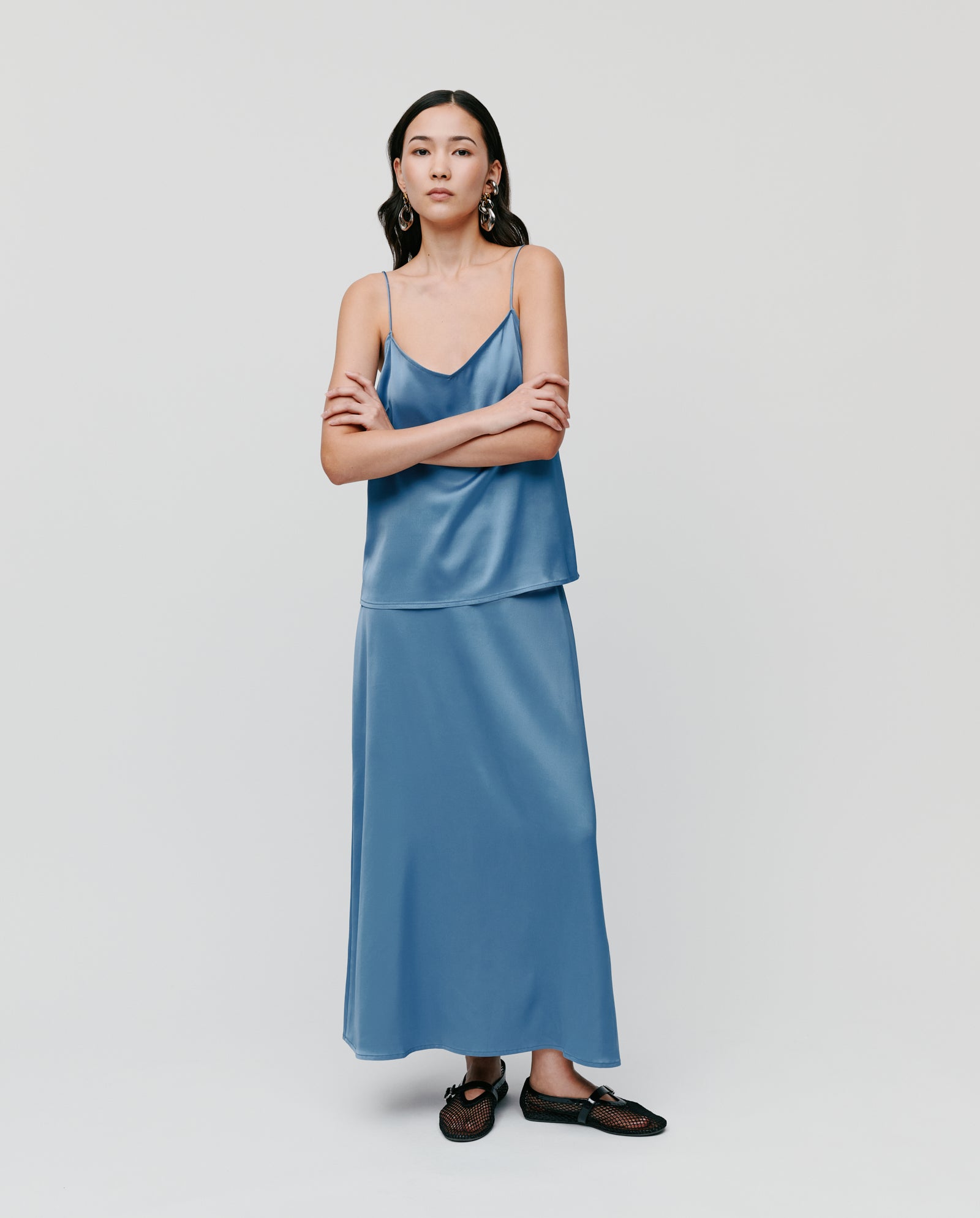 A woman wears the IVY OAK TILDA ANN Straptop, a blue satin V-neck camisole with adjustable straps, paired with a matching long skirt. Made from luxurious Naia acetate, she stands with arms crossed against a plain background.