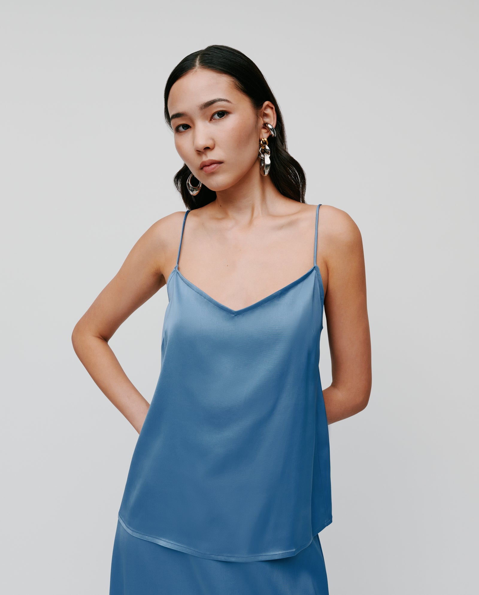 A woman wears the IVY OAK TILDA ANN Straptop in blue satin with a V-neckline and adjustable straps, paired with a skirt and statement earrings against a light background. Made from soft Naia acetate for an elegant look.