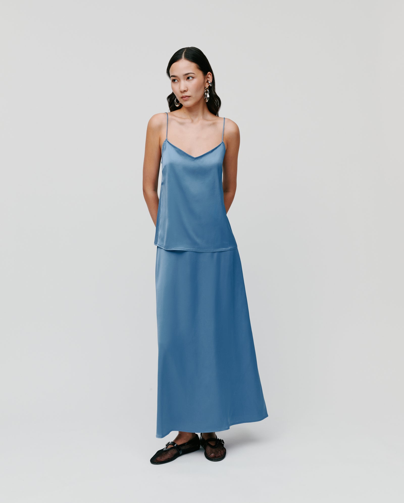A woman in the IVY OAK TILDA ANN Straptop, a blue satin V-neck camisole with adjustable straps, and a matching Naia acetate skirt, paired with black shoes, stands against a plain background.