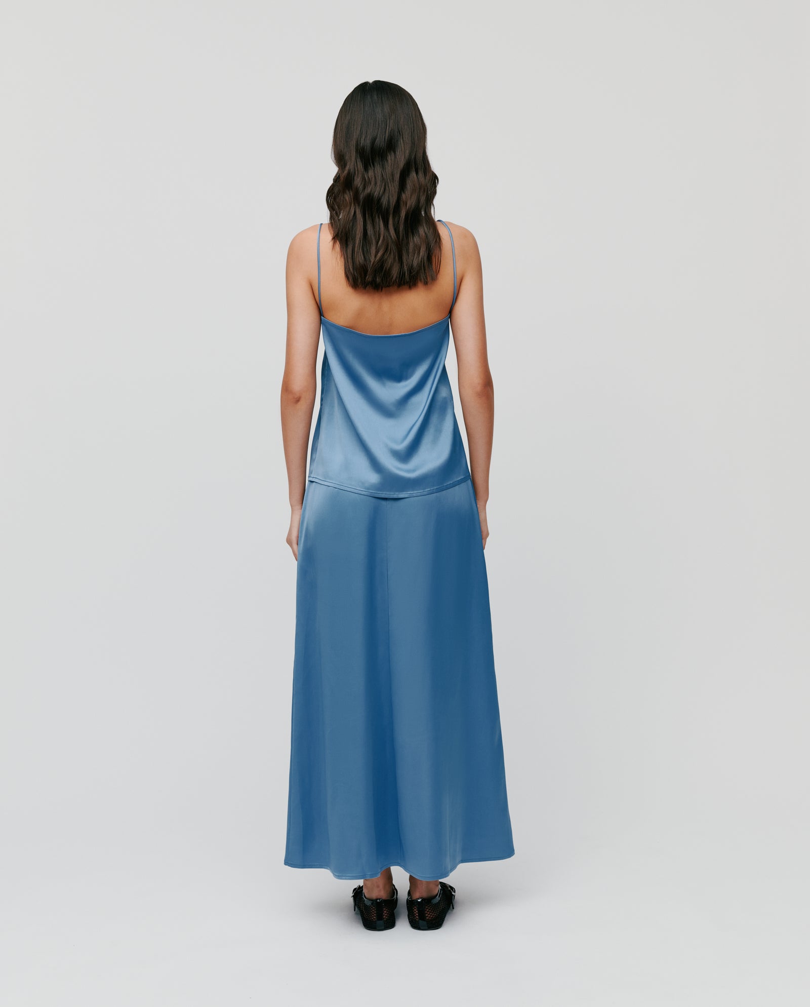 Woman with wavy brown hair wearing the IVY OAK TILDA ANN Straptop in blue satin, featuring adjustable straps and a V-neckline made from sustainable Naia acetate, plus black shoes, facing away from the camera.