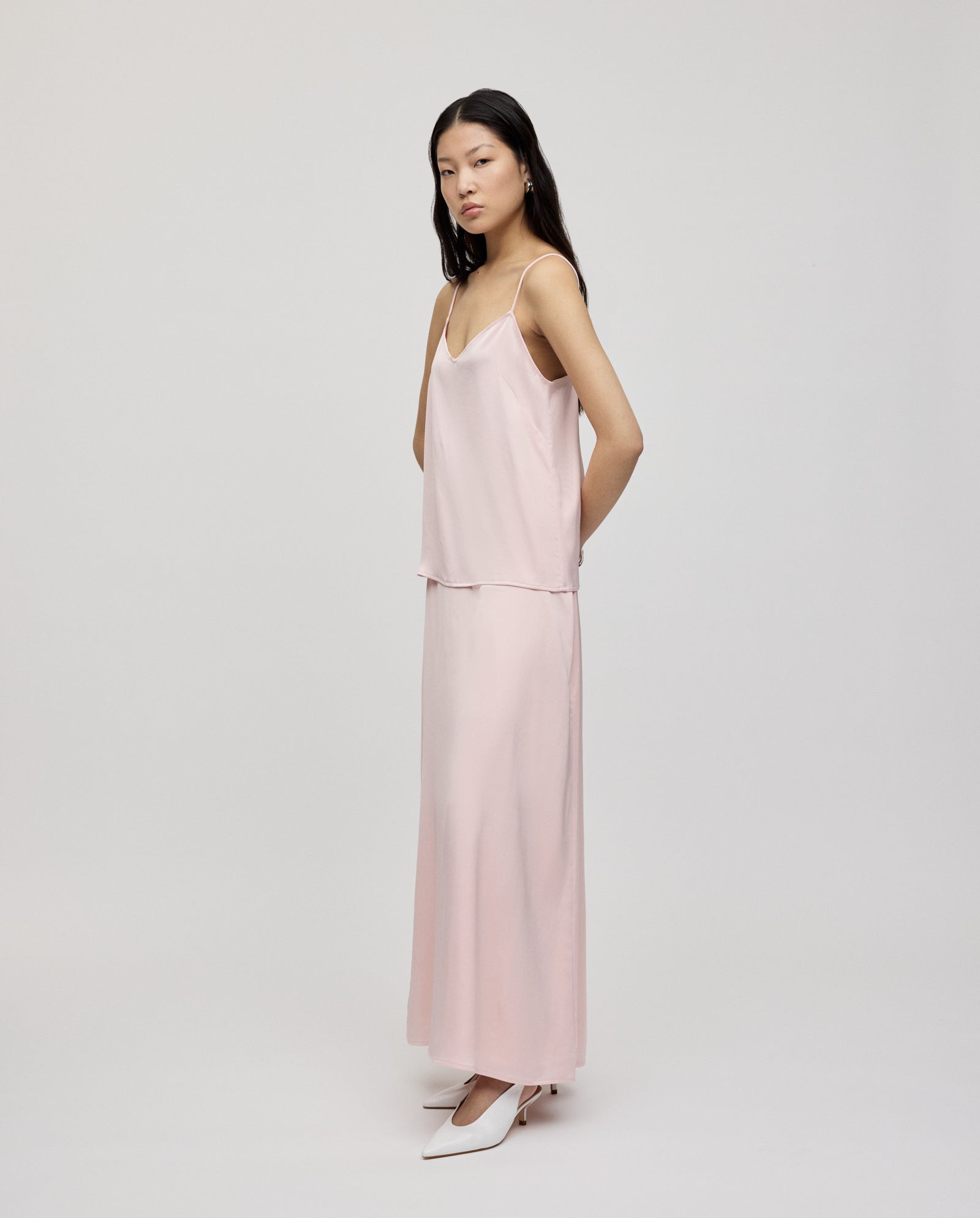 Woman in a light pink IVY OAK TILDA ANN Straptop with a V-neckline and long skirt, crafted from silky Naia acetate, paired with white heels, standing against a plain background.