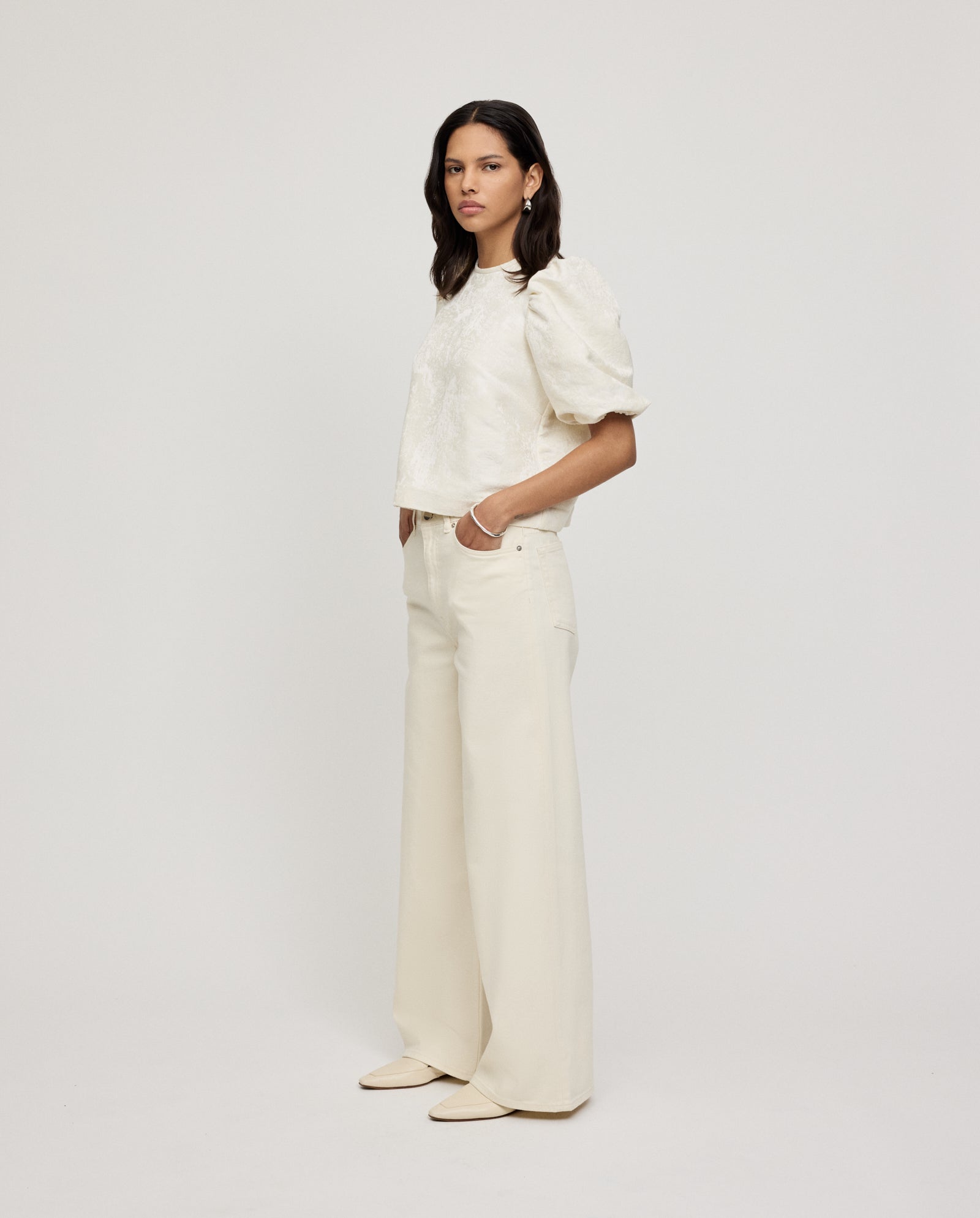 A woman wears the IVY OAK ELISABETH Top, a jacquard puff-sleeve white blouse, paired with wide-leg cream pants, standing against a plain light background.
