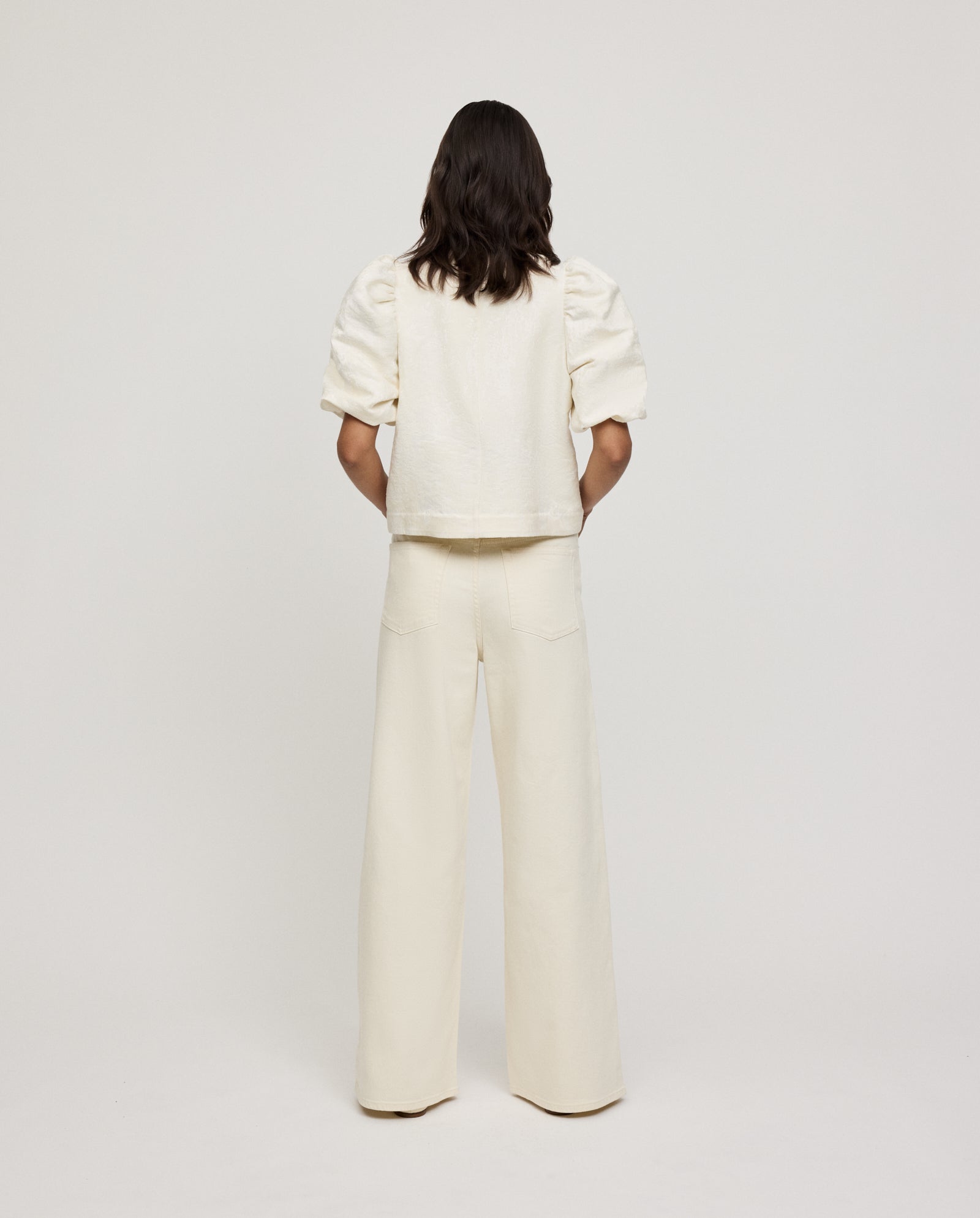 A person with long hair is seen from behind wearing the IVY OAK ELISABETH Top, a cream jacquard blouse, paired with wide-leg pants.