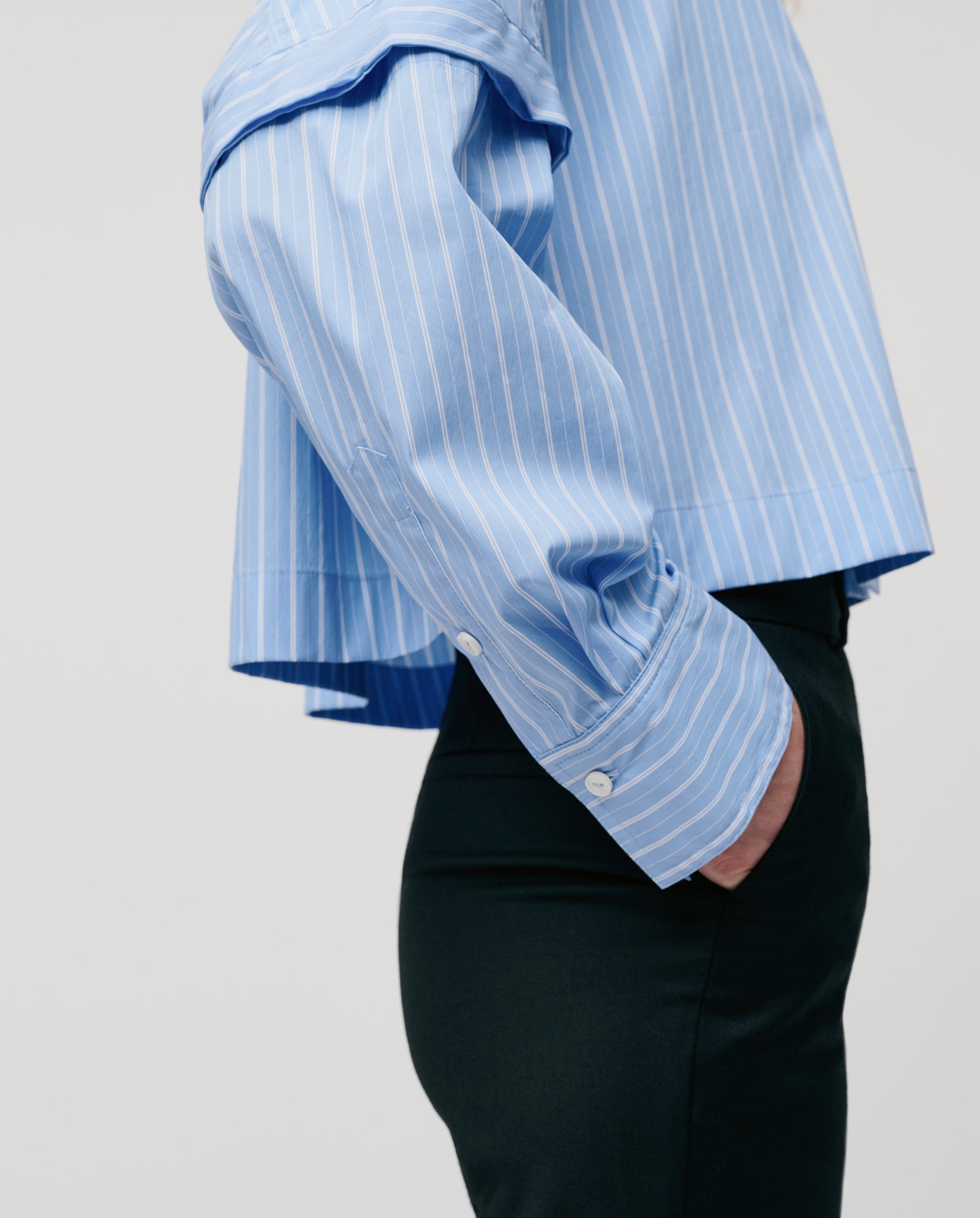 Model in ELVIRA Blouse by IVY OAK, a blue striped cropped shirt, paired with black pants. Seen from the side with one hand in pocket.