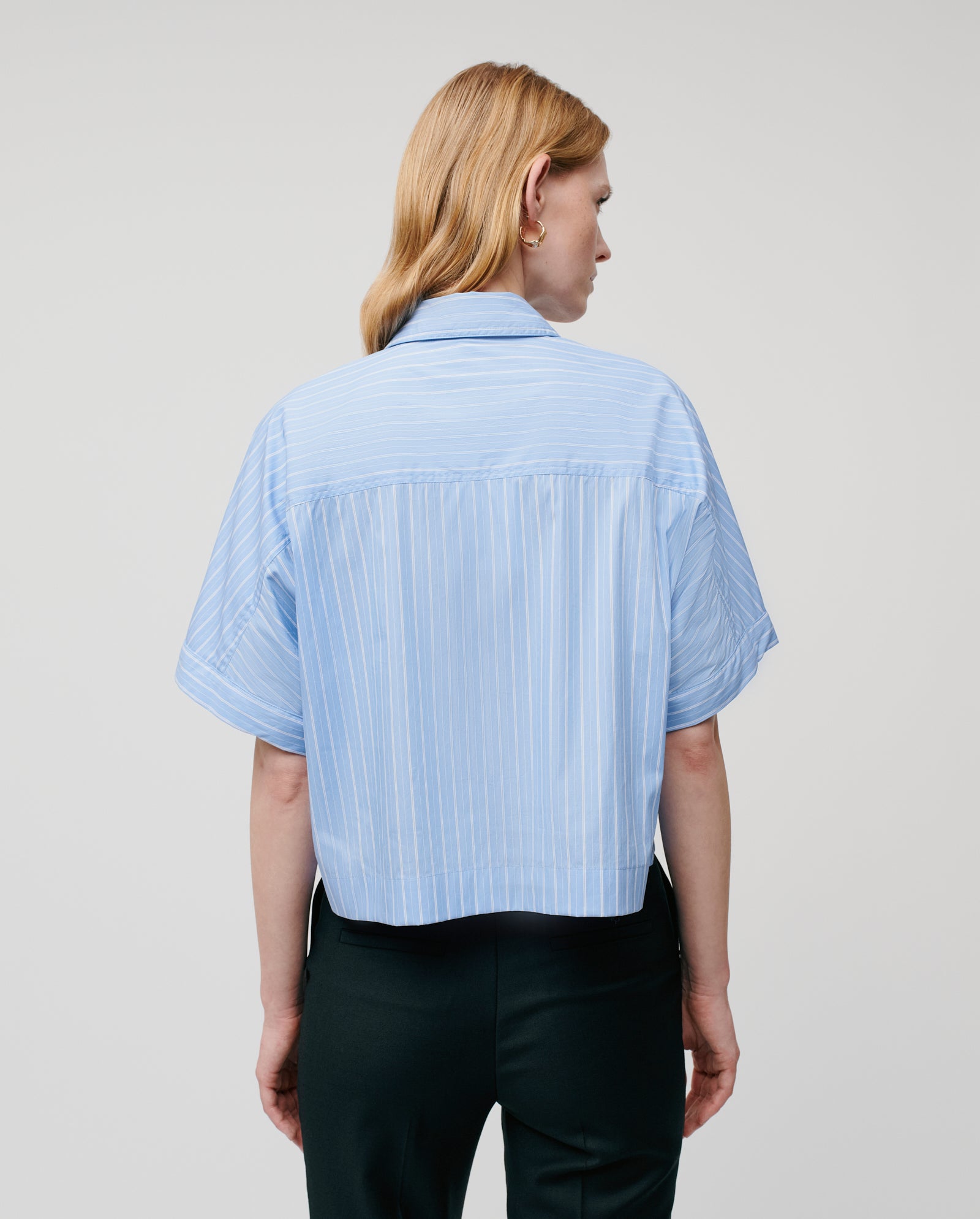 A person with light hair is seen from behind against a plain background, wearing the IVY OAK ELVIRA Blouse—a light blue cropped shirt with detachable sleeves.