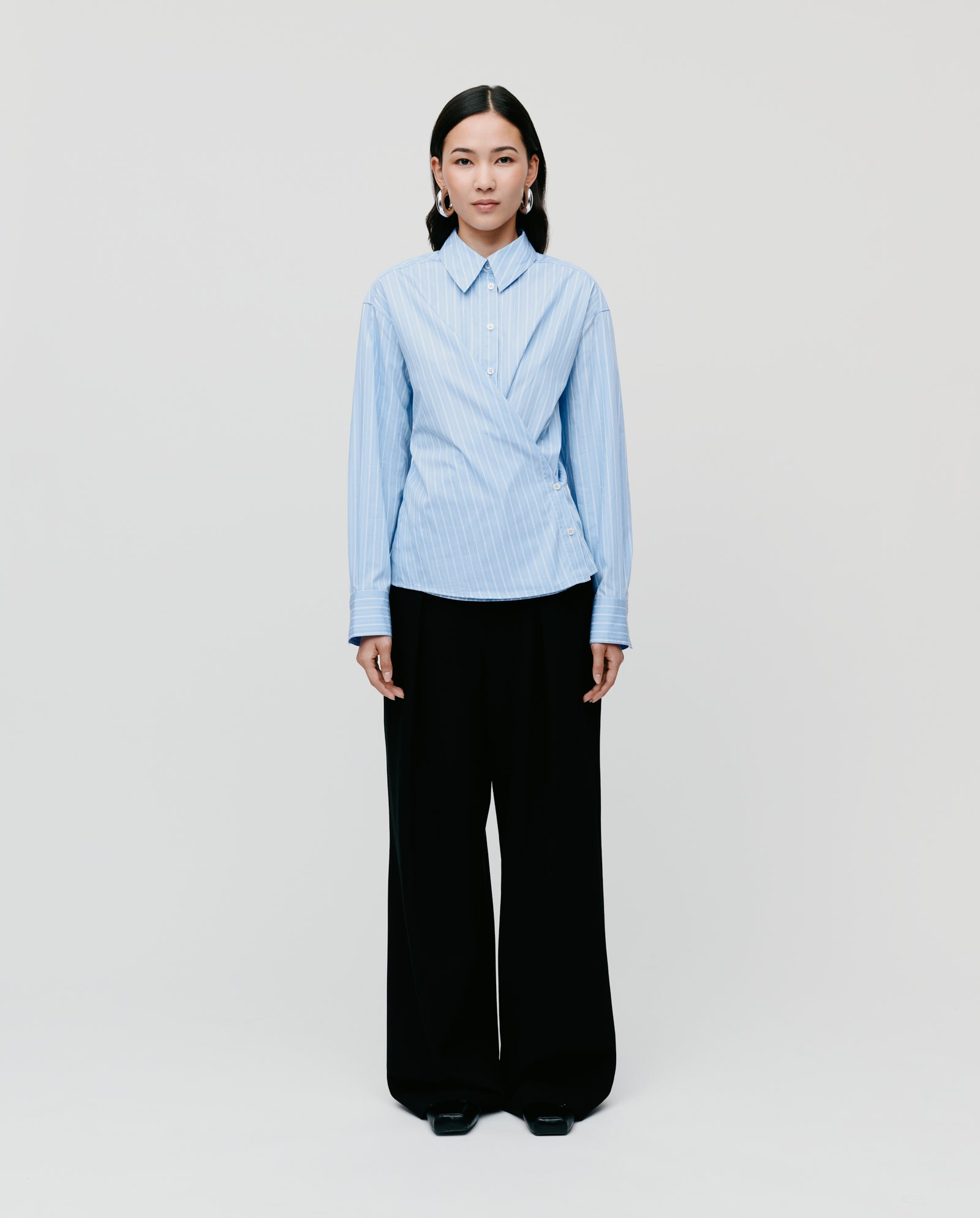 A woman faces forward in the IVY OAK EVONNE Shirt, an oversized blue blouse with a button placket, paired with wide-leg black pants against a plain background.