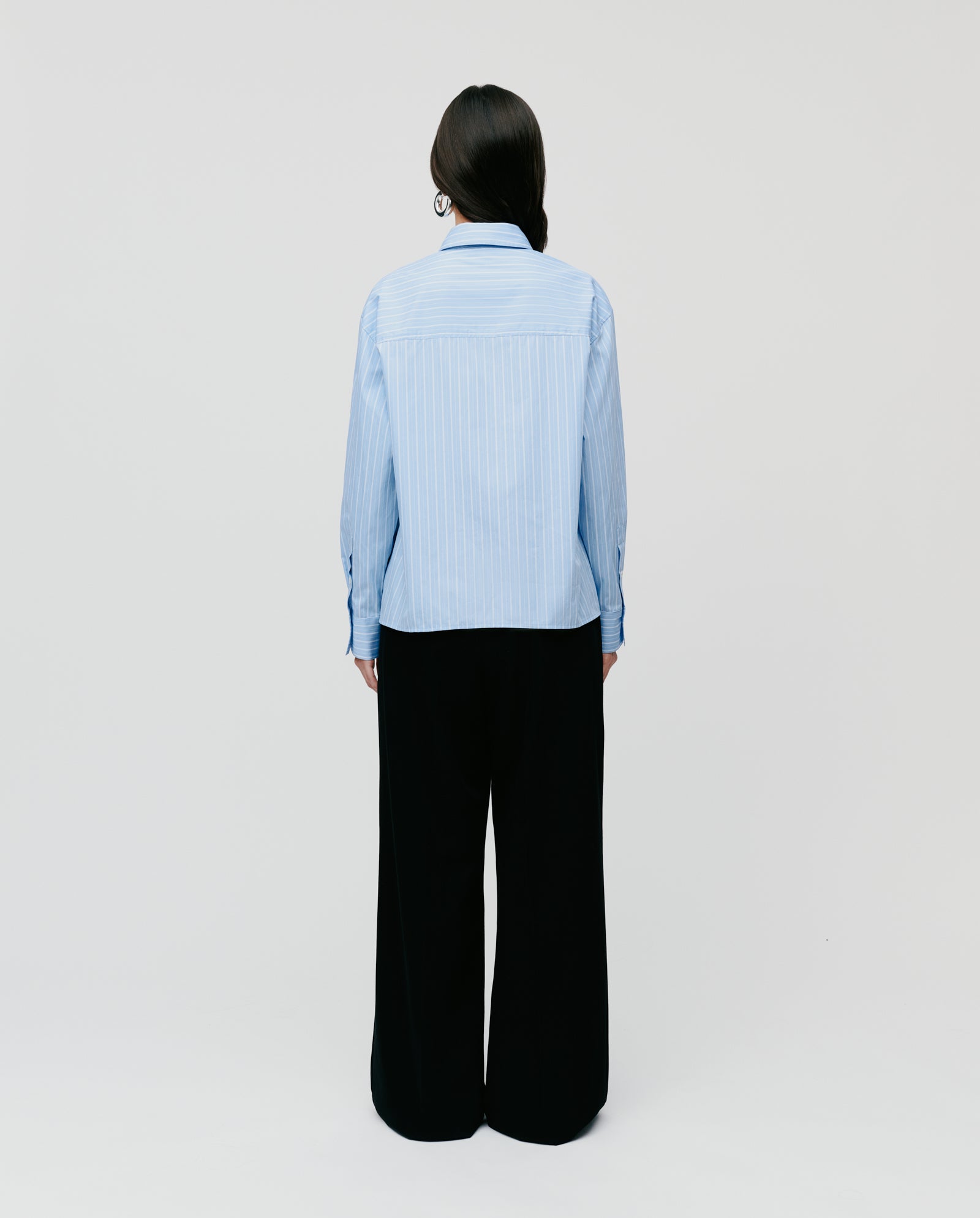 A person stands facing away, wearing the oversized EVONNE Shirt by IVY OAK with light blue stripes and loose black pants against a plain background.