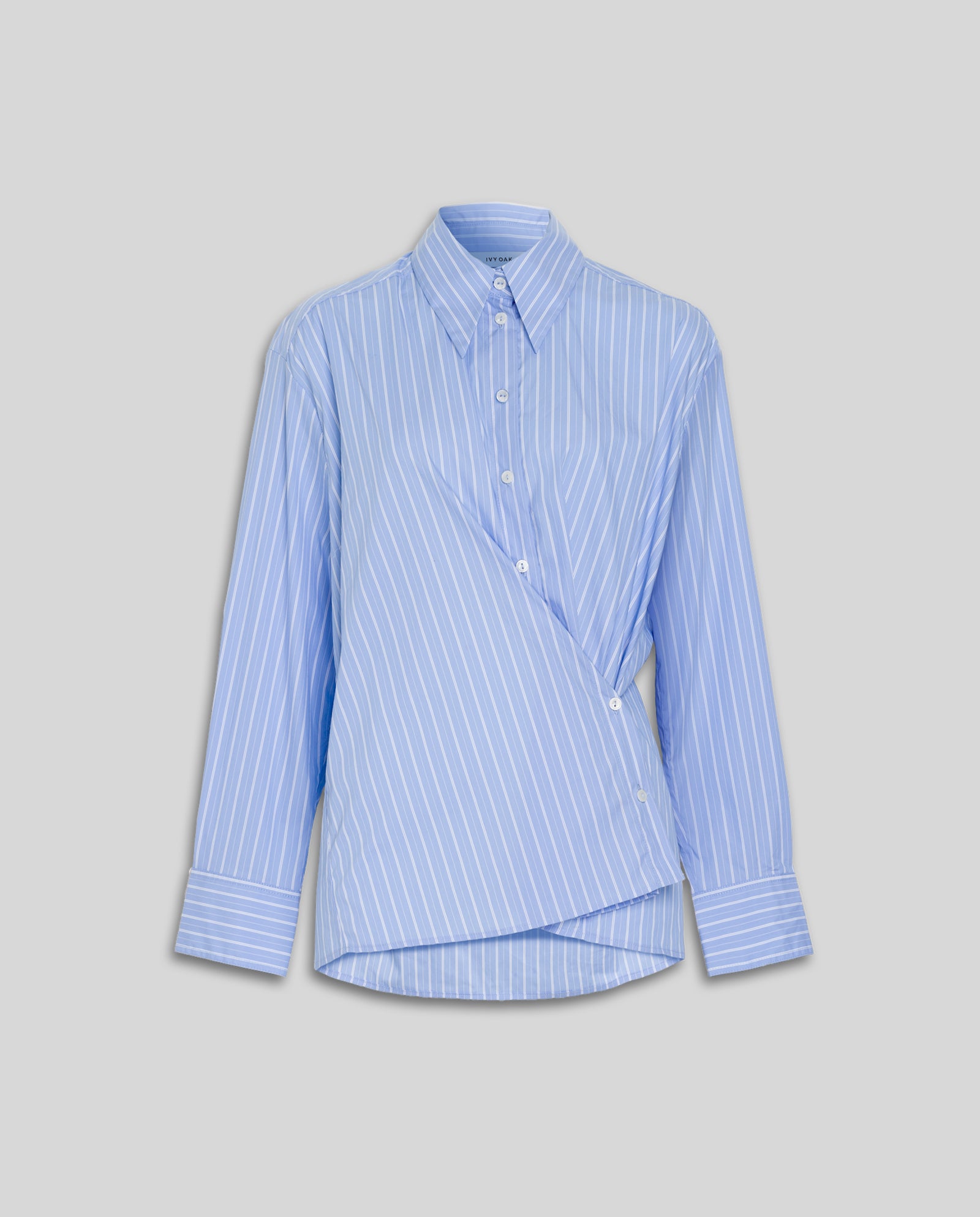 The IVY OAK EVONNE Shirt is a light blue oversized blouse with white vertical stripes, an asymmetric twisted front, and a chic button placket, shown on a gray background.