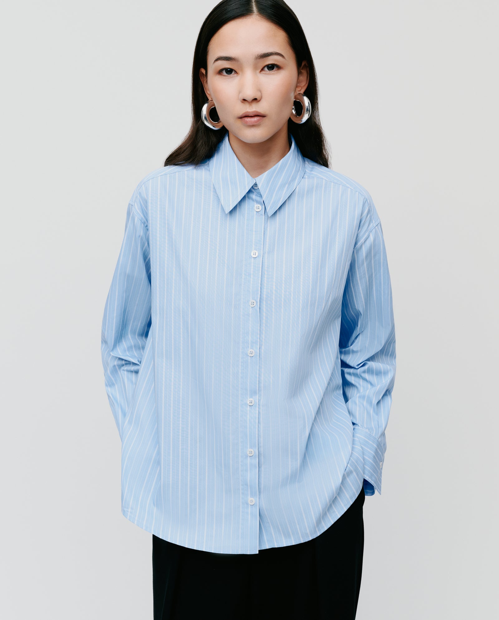 Woman wearing the IVY OAK EVONNE Shirt, a light blue striped oversized blouse with button placket, hands in pockets, standing against a plain background.
