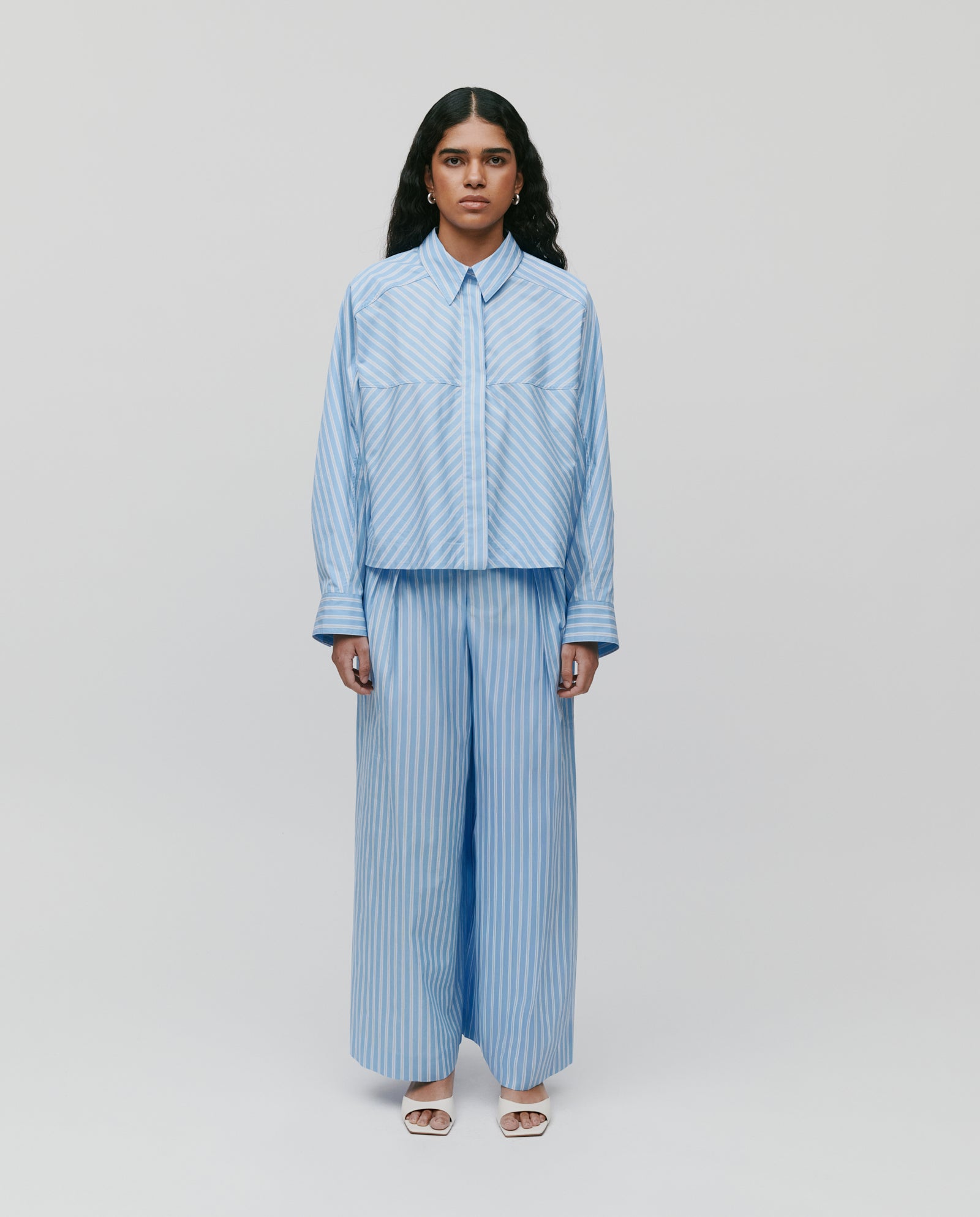 A person stands facing forward, wearing the IVY OAK BRITTNEY Blouse— a boxy, loose light blue striped shirt—paired with matching wide-leg pants.