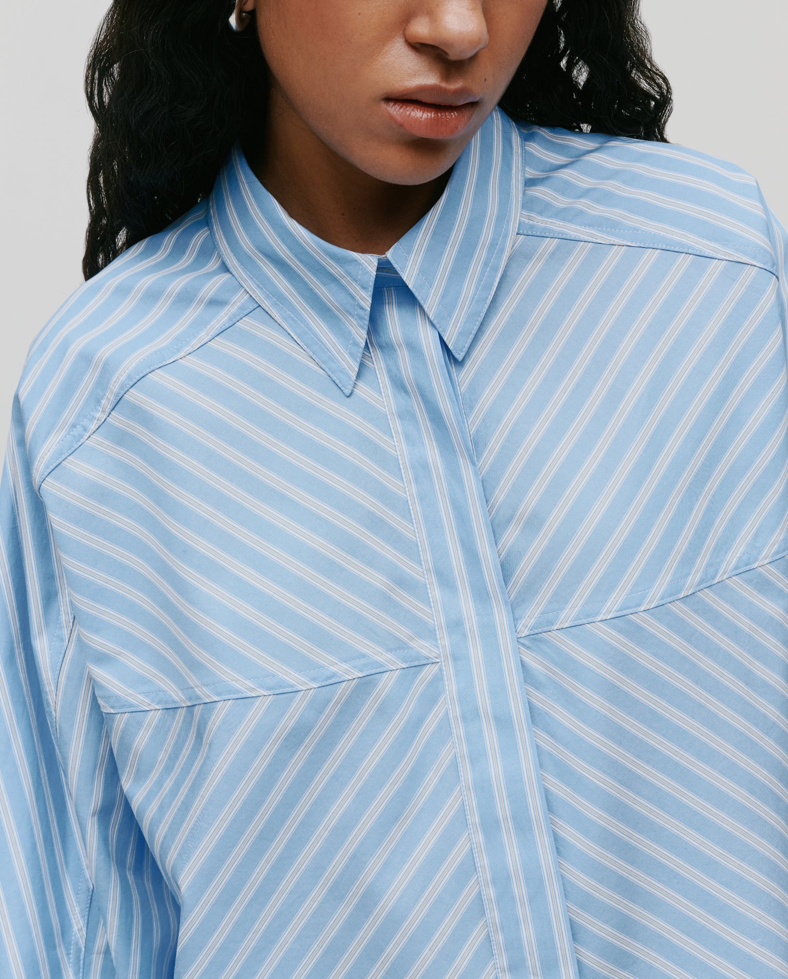 A woman wears the IVY OAK BRITTNEY Blouse, a boxy-fit button-up shirt with light blue and white stripes, shown from the shoulders up.