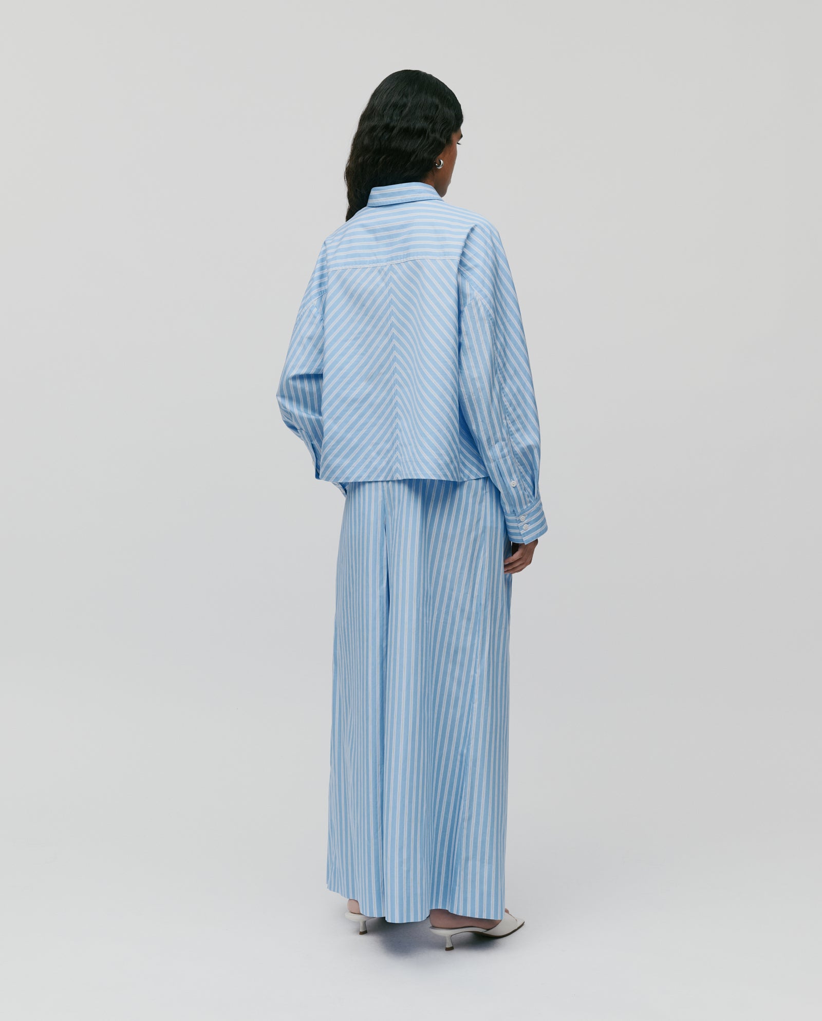 Model wears IVY OAKs BRITTNEY Blouse in a boxy fit, light blue and white striped design with stripe inlay, paired with matching wide-leg pants, shown from the back.