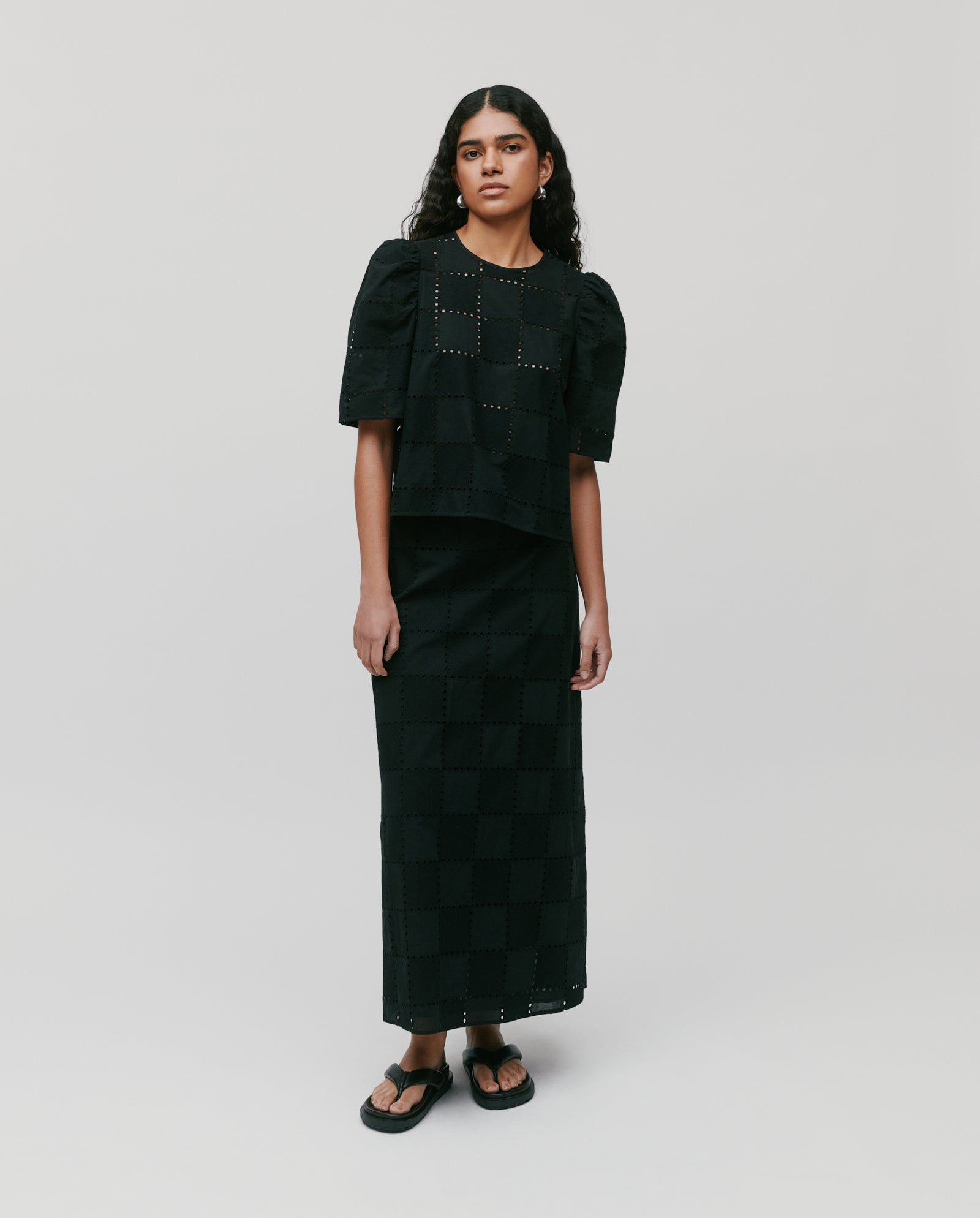 A woman wears the IVY OAK ELISABETH ANN Top, a black broderie anglaise blouse with gathered sleeves, paired with a long checkered skirt and stands against a plain background.