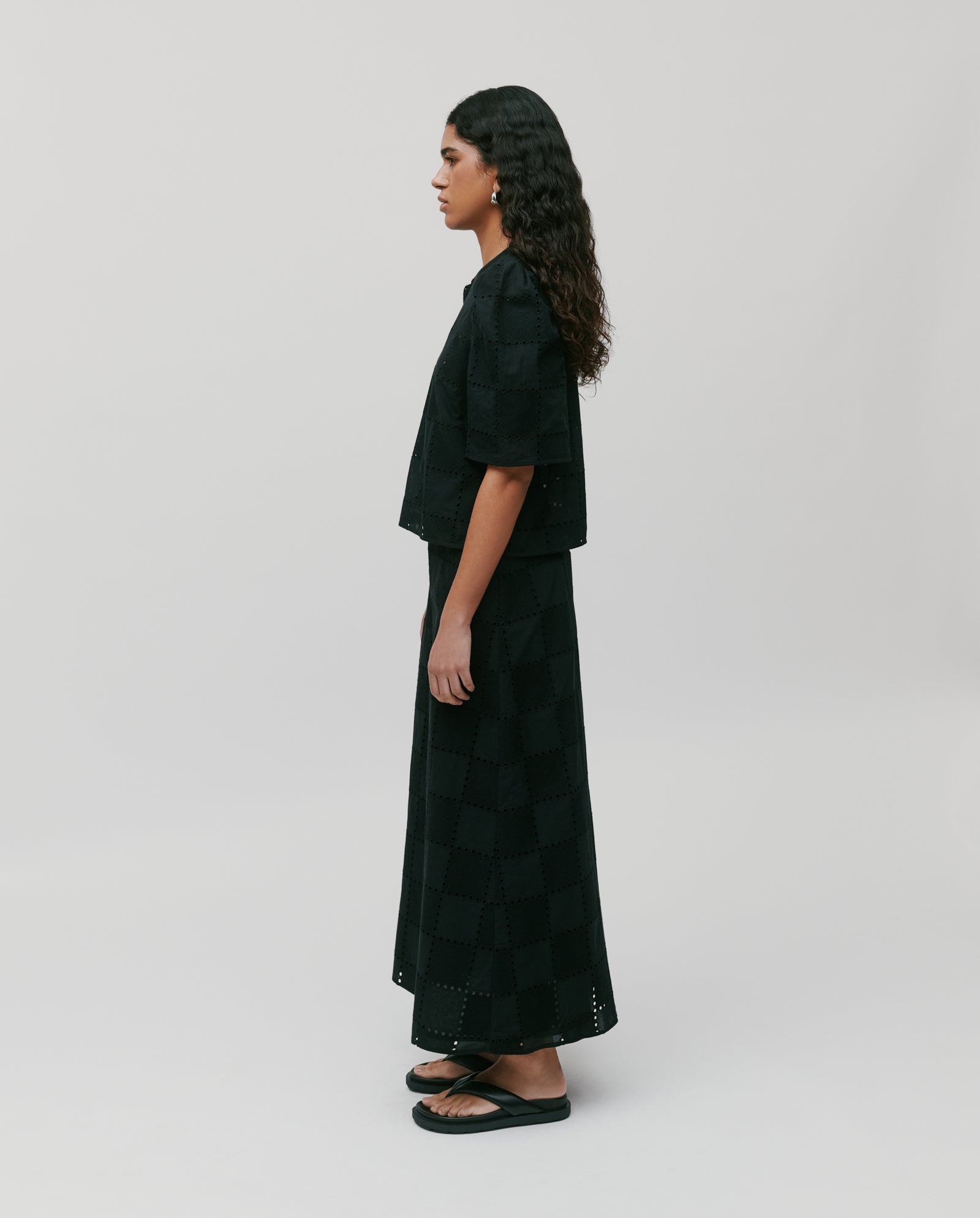 Woman in the IVY OAK ELISABETH ANN Top—a black broderie anglaise blouse with gathered sleeves—paired with a long skirt and sandals, standing sideways against a plain background.