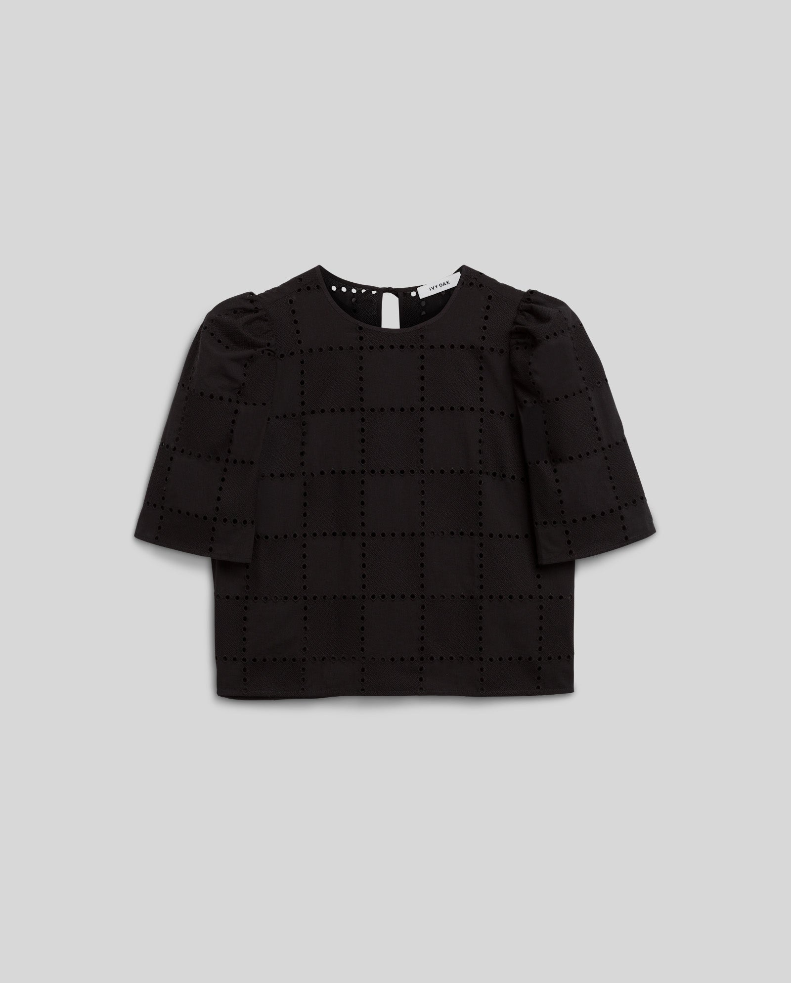 The IVY OAK ELISABETH ANN Top is a black short-sleeve blouse featuring a grid broderie anglaise pattern and back button closure, shown against a grey background.