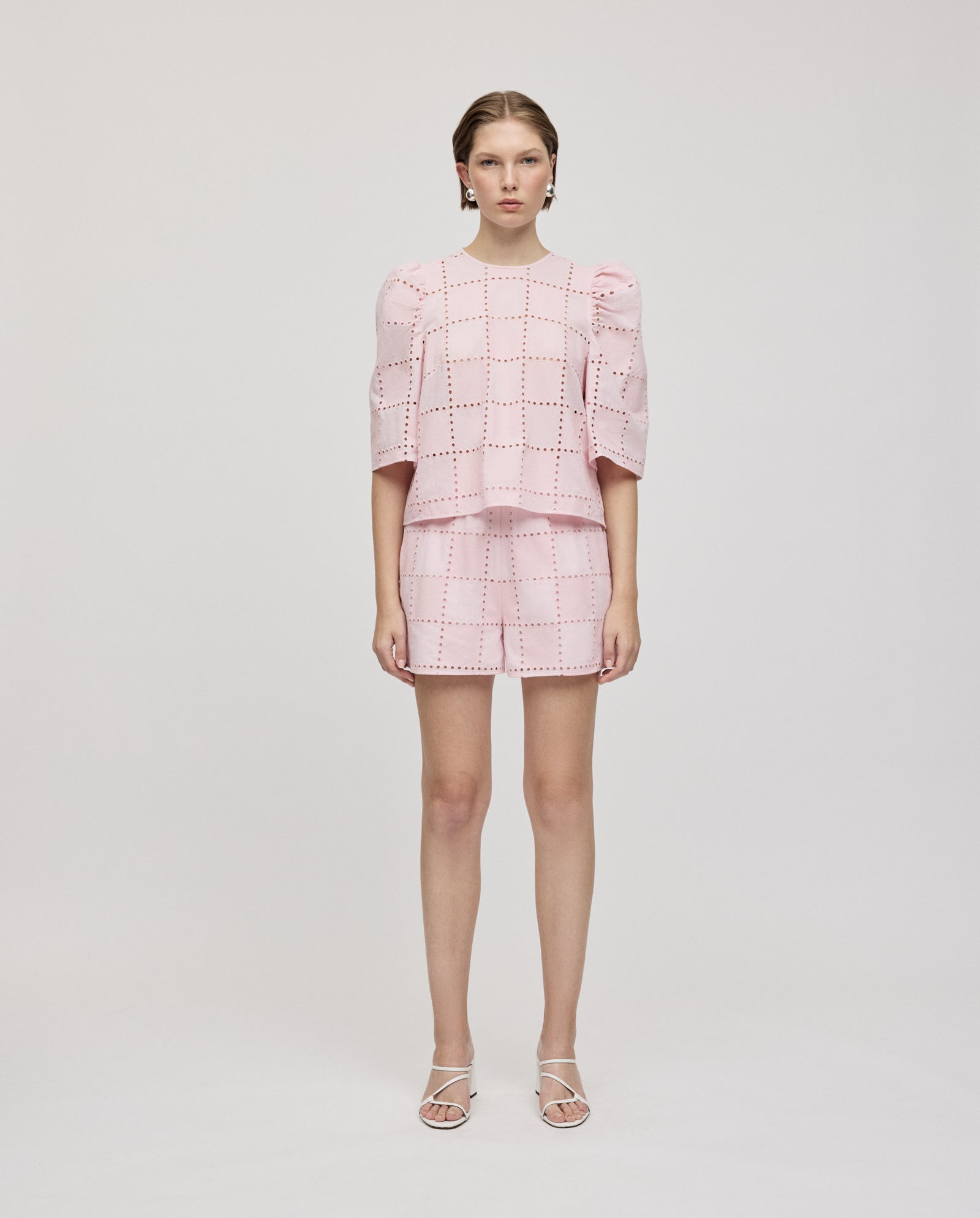 A woman wears the ELISABETH ANN Top by IVY OAK, a light pink broderie anglaise style with square cutout patterns and gathered sleeves, paired with matching shorts against a plain background.