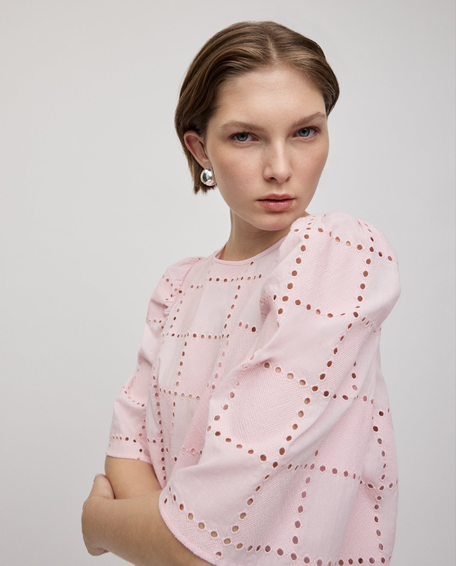 Woman wearing the ELISABETH ANN Top by IVY OAK in pink broderie anglaise with gathered sleeves, arms crossed and looking seriously at the camera.