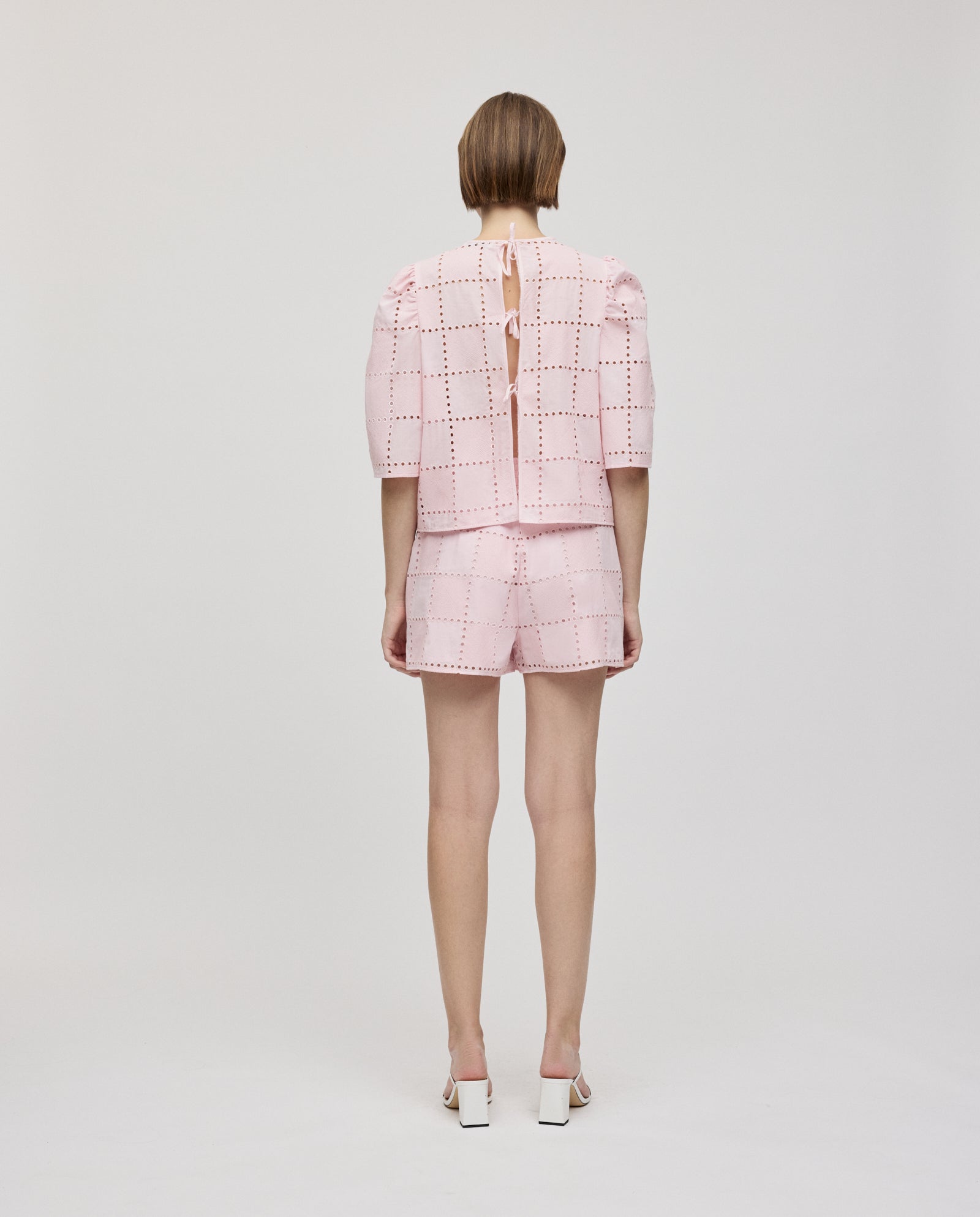 A person stands facing away, wearing the IVY OAK ELISABETH ANN Top in a pink grid pattern with gathered sleeves, paired with matching shorts and heeled sandals.