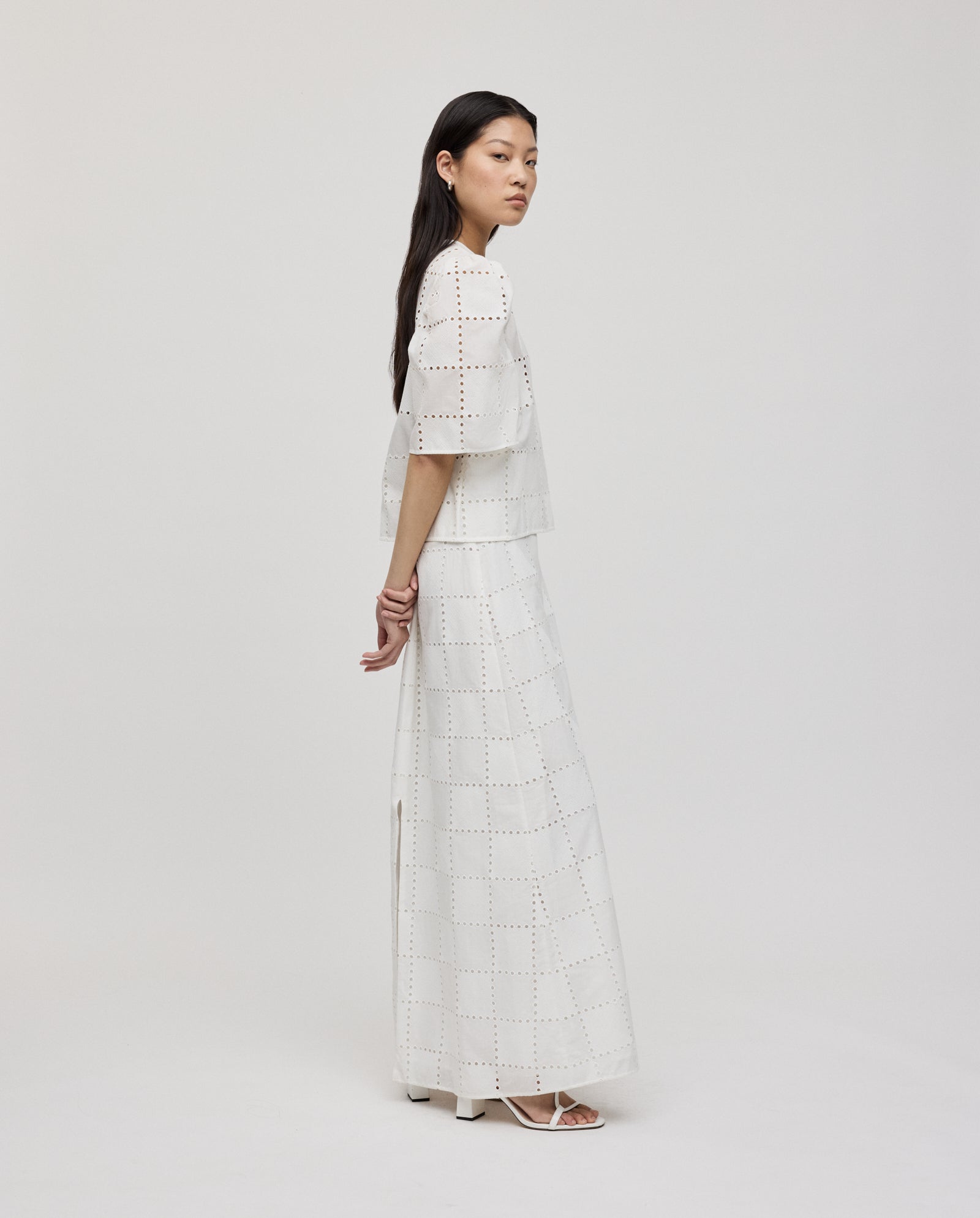A woman stands sideways against a plain background, wearing the IVY OAK ELISABETH ANN Top—a white, grid-patterned broderie anglaise blouse with gathered sleeves—paired with a long skirt.