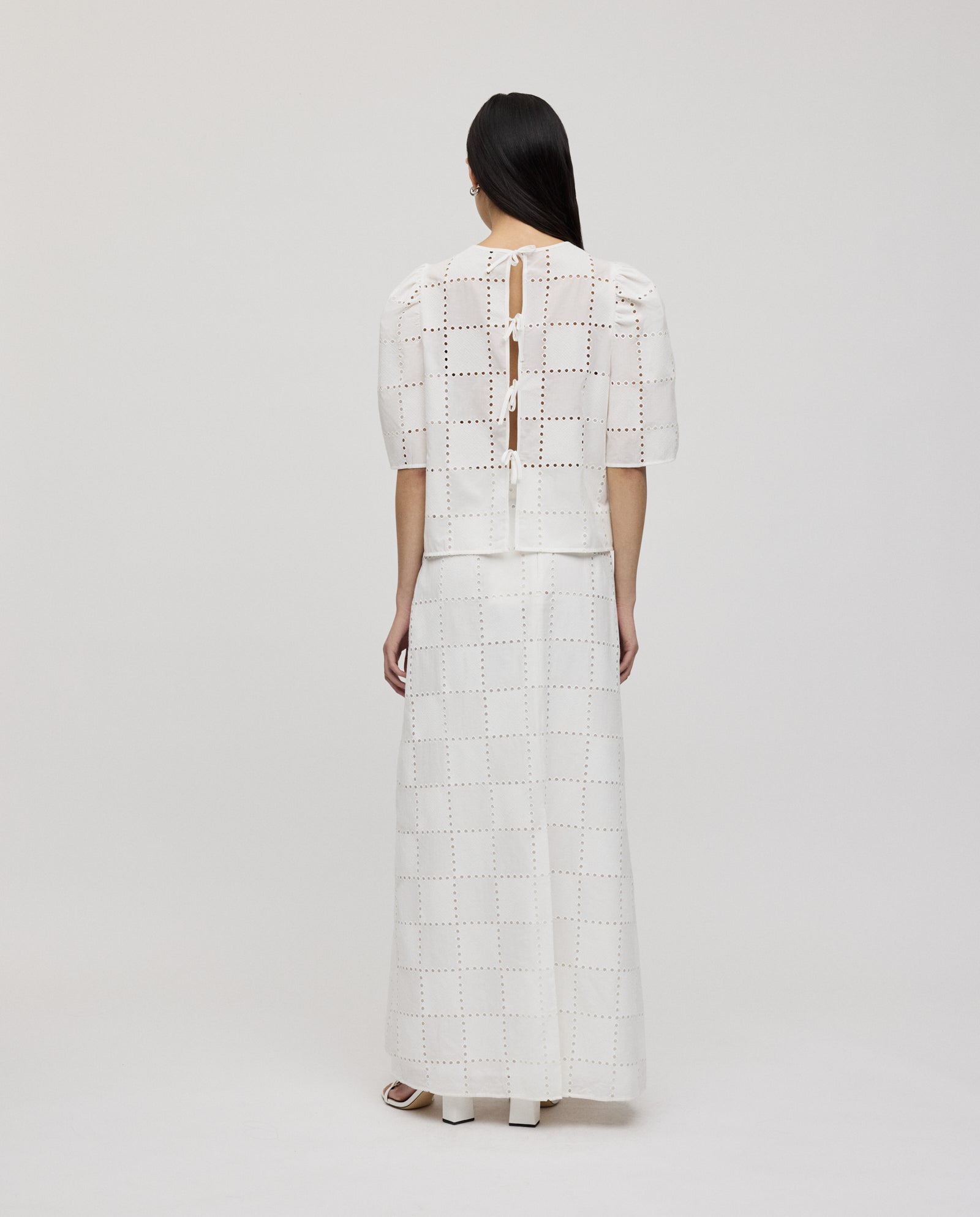 A woman stands with her back to the camera, wearing the IVY OAK ELISABETH ANN Top—a white broderie anglaise blouse with gathered sleeves—and a long skirt.