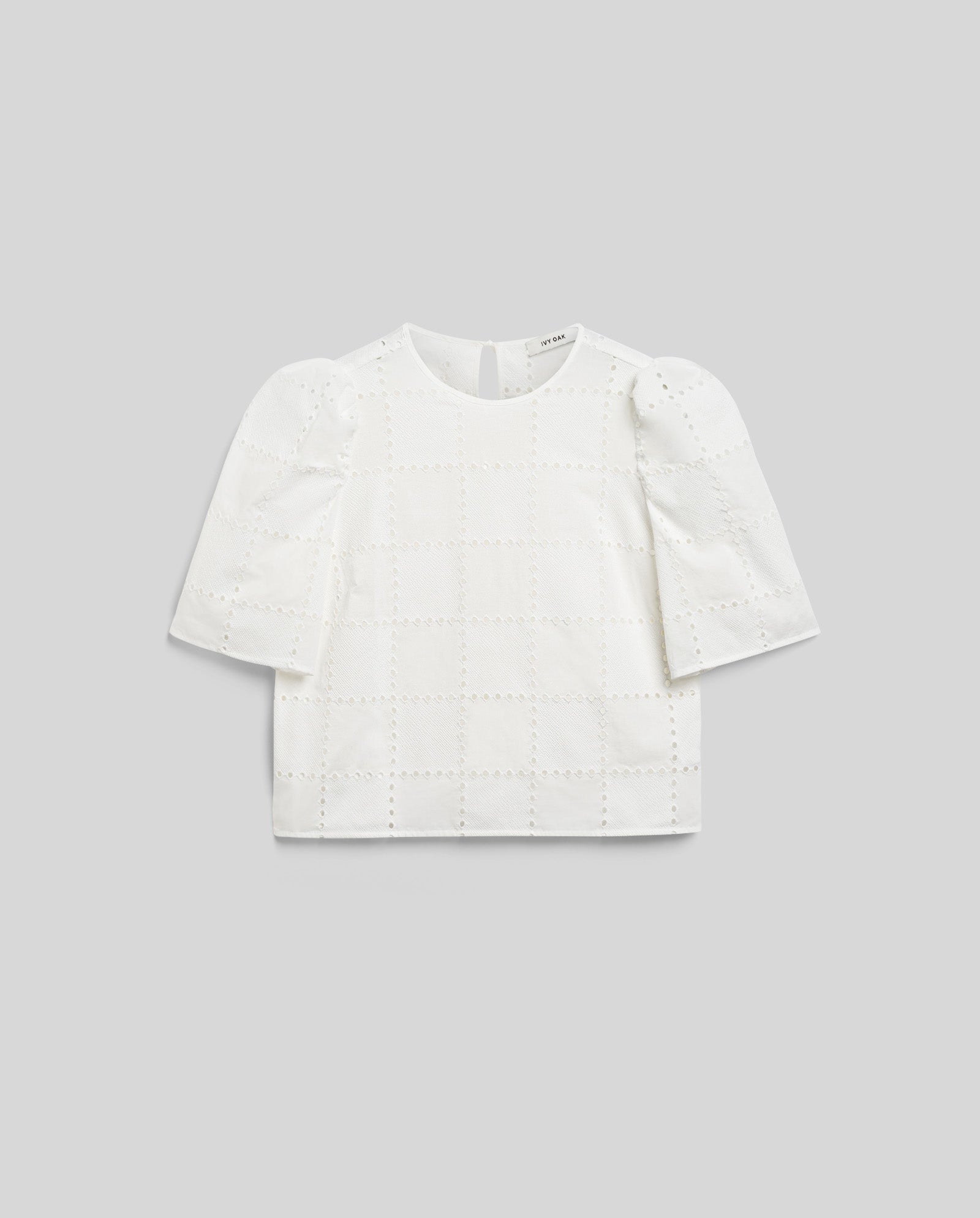 The ELISABETH ANN Top by IVY OAK is a white, short-sleeved blouse with gathered sleeves, puffed shoulders, and a subtle broderie anglaise checkered eyelet pattern, shown against a gray background.