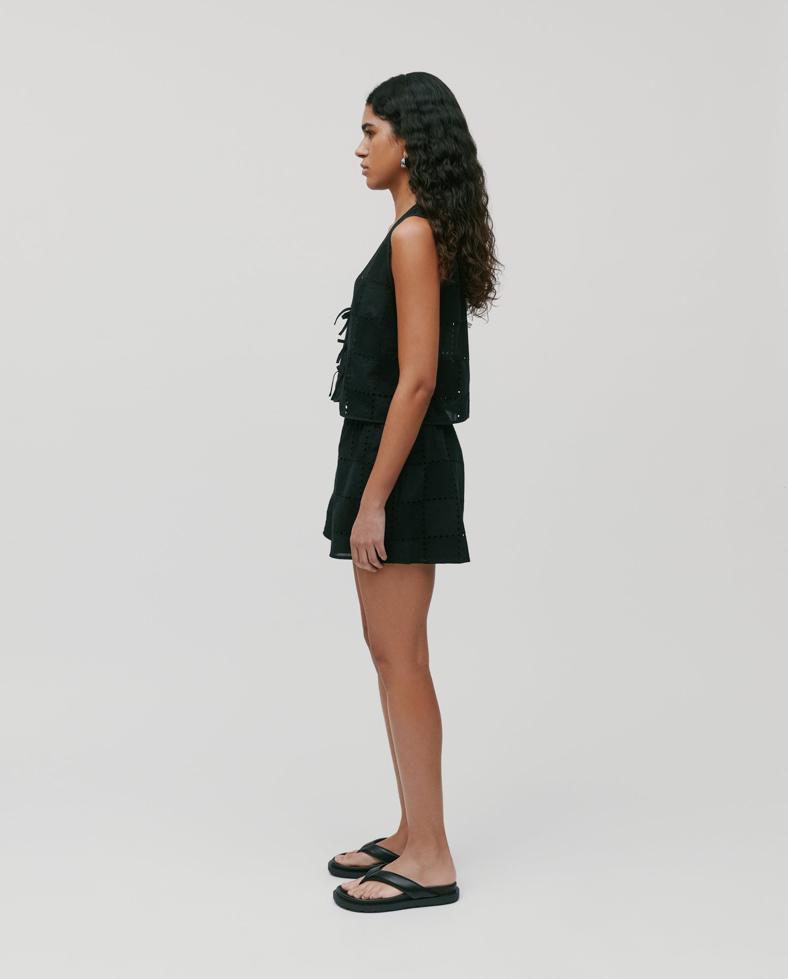 A woman with long curly hair wears the IVY OAK TALA Top, paired with shorts and sandals, standing in profile against a white background.