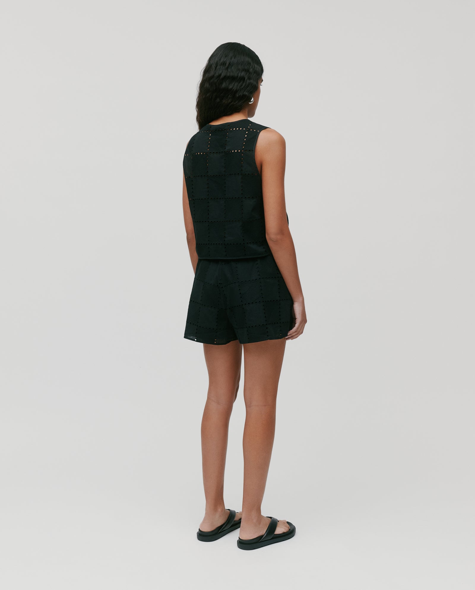 Person standing and facing away, wearing a black sleeveless IVY OAK TALA Top, black shorts, and black sandals.