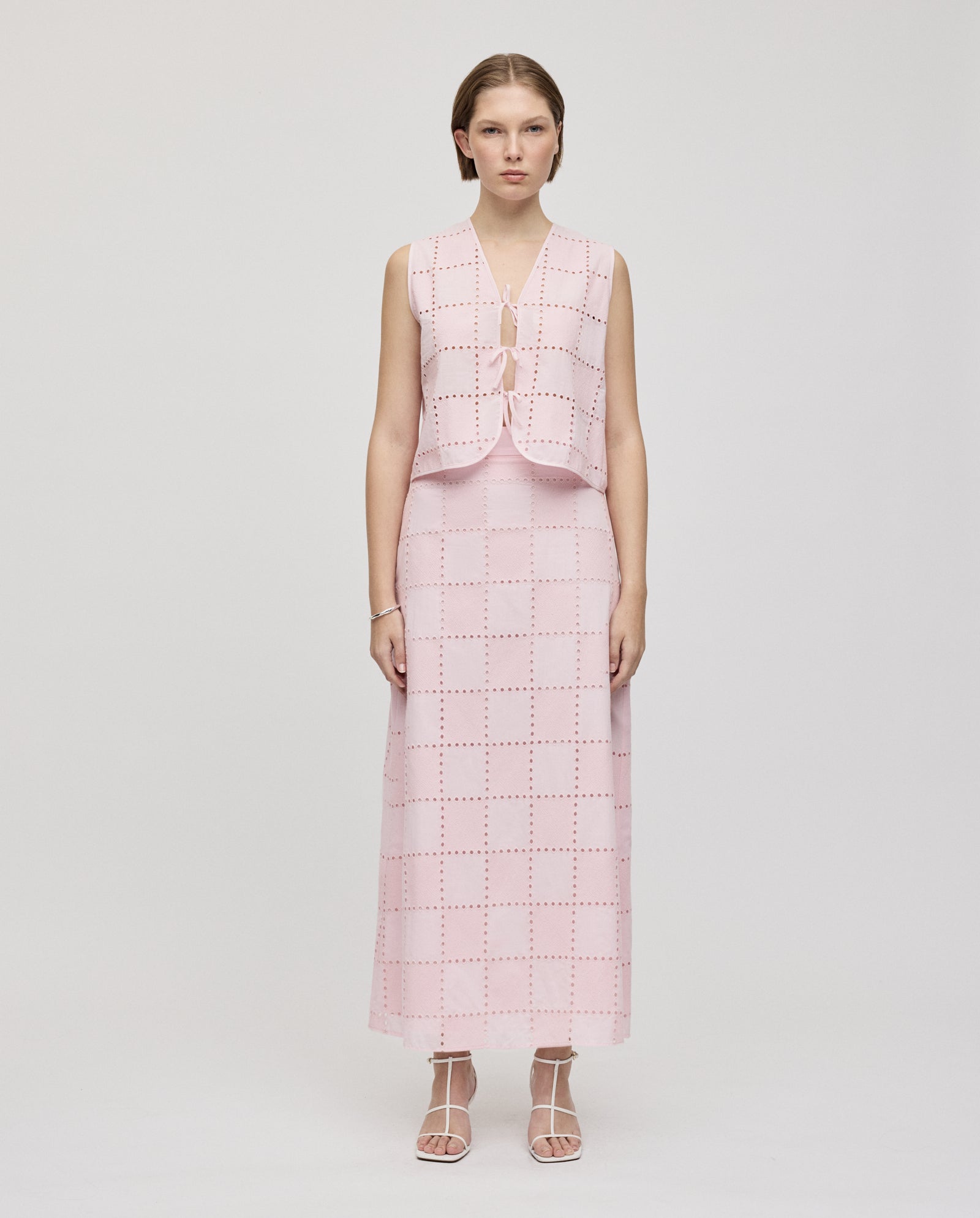 A woman stands straight in the IVY OAK TALA Top, a sleeveless, light pink checkered piece with delicate embroidered anglaise details, paired with a matching vest and strappy sandals for an elevated look.