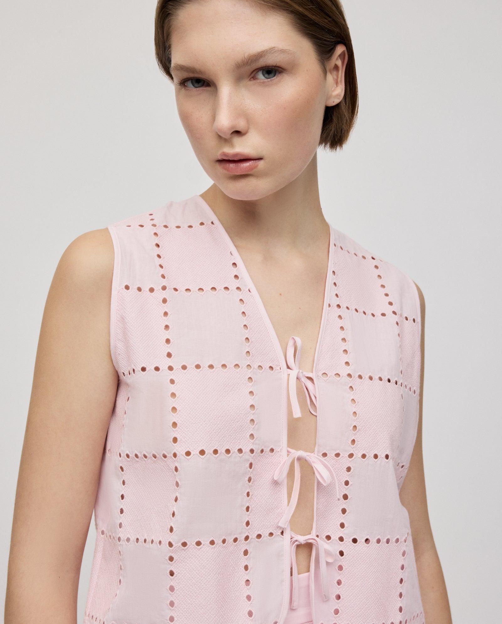 A woman models the IVY OAK TALA Top, a sleeveless light pink blouse featuring embroidered anglaise square cutout patterns and front tie closures, while looking at the camera.