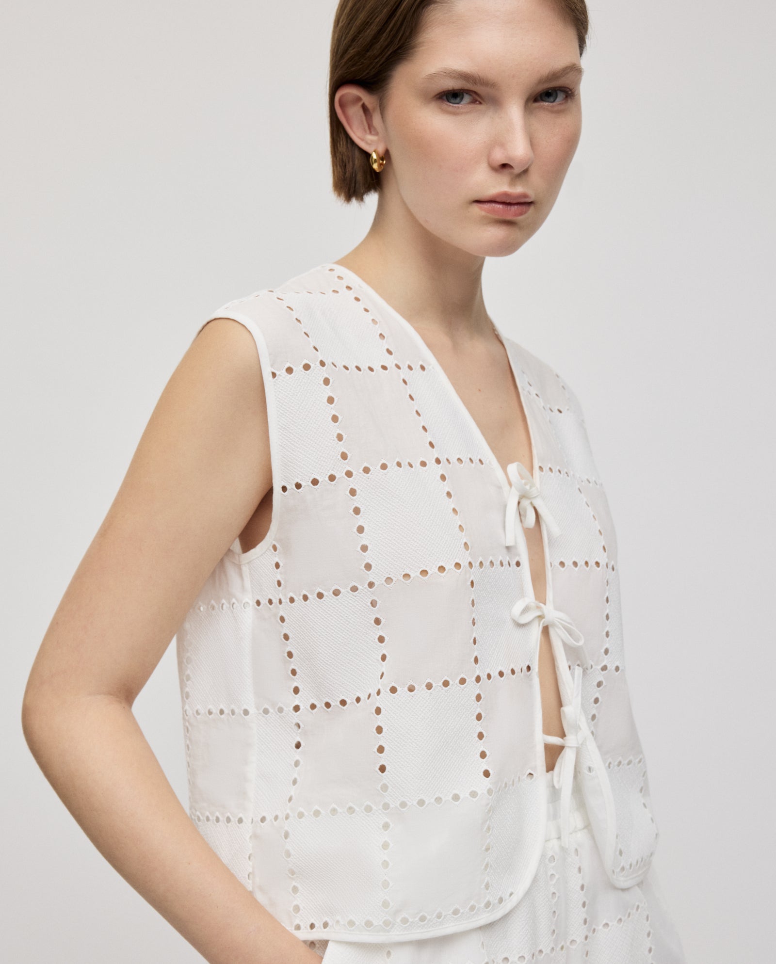 A woman wears the IVY OAK TALA Top, a white sleeveless piece with delicate embroidered anglaise and bow ties, set against a plain background.