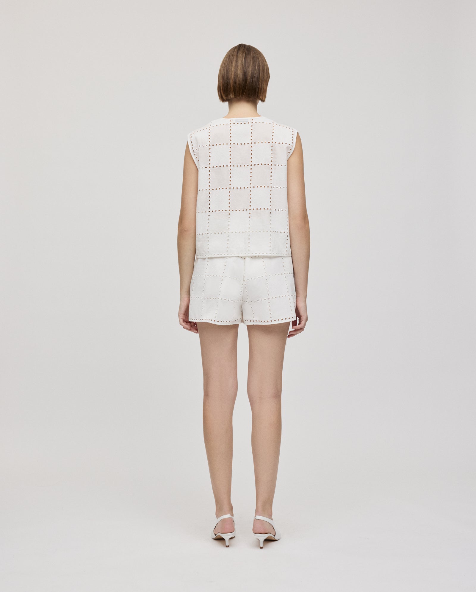 A woman faces away, wearing an IVY OAK TALA Top in white and matching shorts with square cutout details, paired with heels.