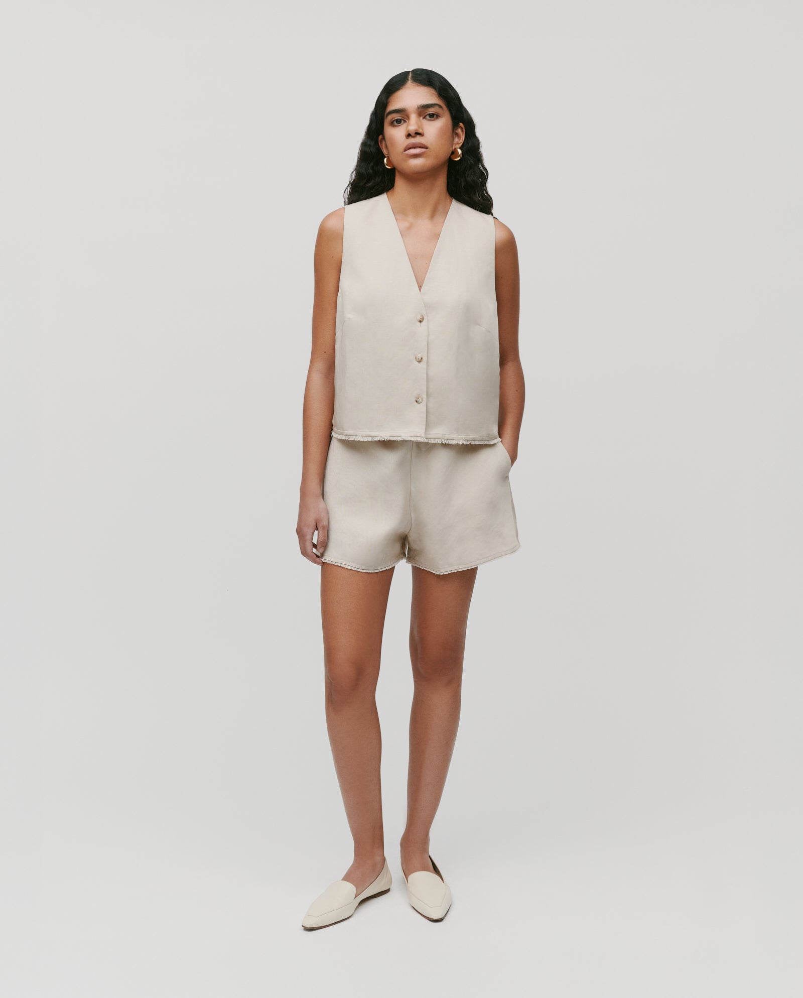 A woman wears the IVY OAK TALA ANN Top—a beige sleeveless linen-blend vest with front button closure—paired with matching shorts and beige flats, standing against a plain background.