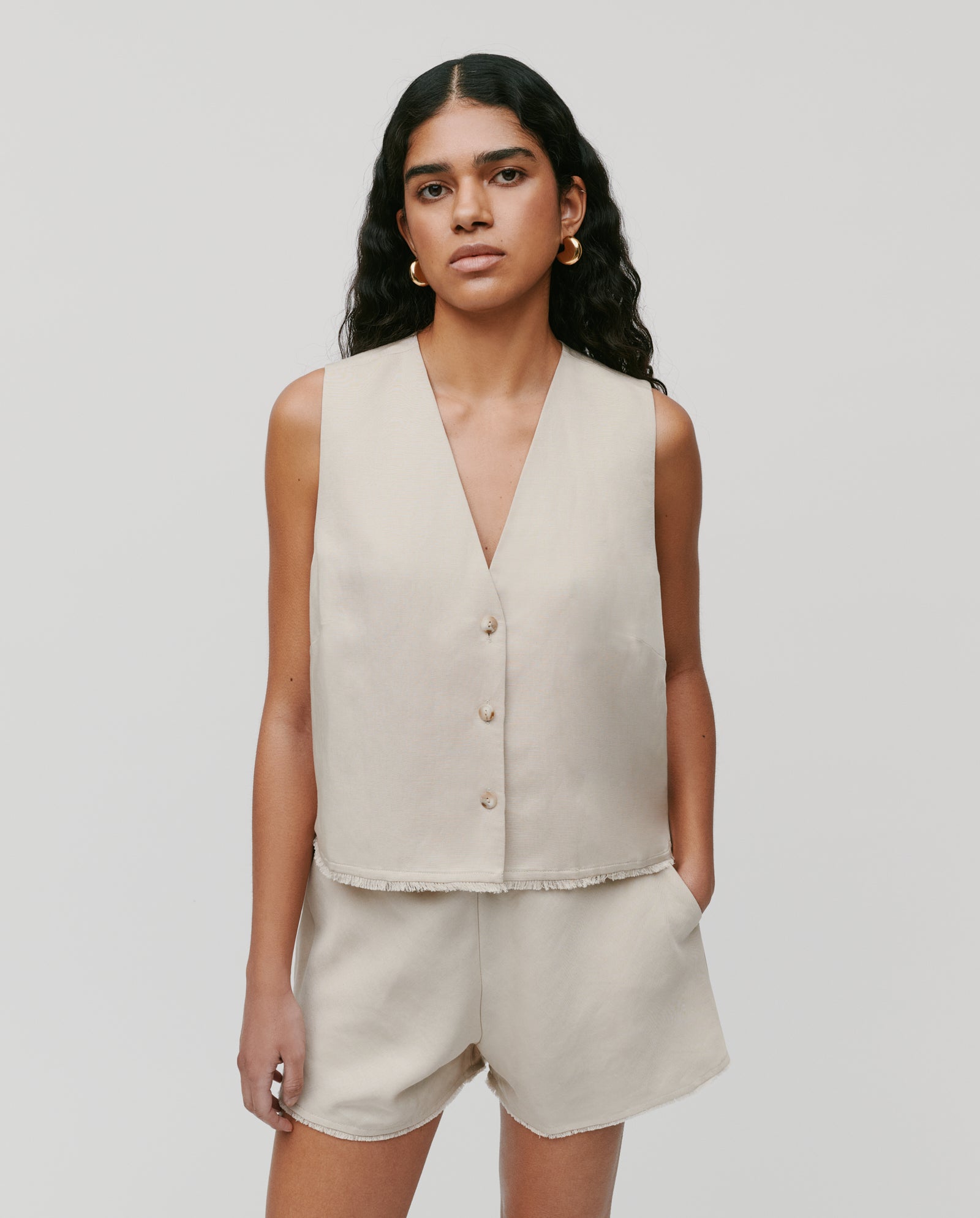 A woman wears the IVY OAK TALA ANN Top, a beige linen-blend sleeveless vest with front buttons, paired with matching shorts, standing against a plain light background.