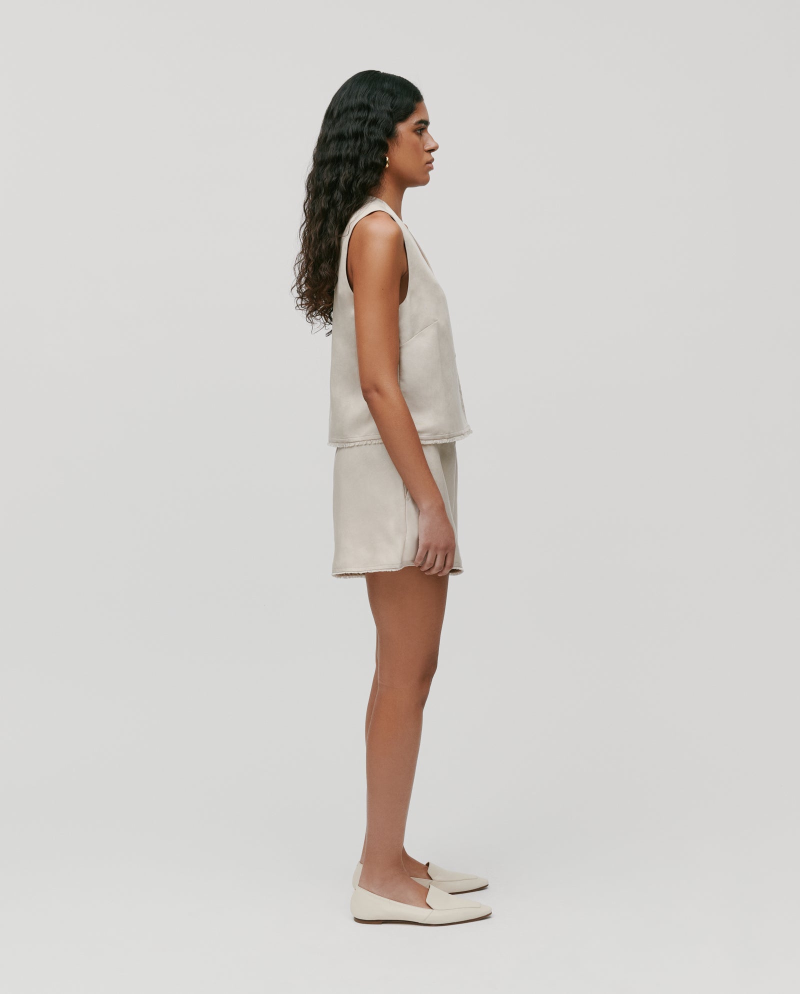 A woman stands in profile, wearing the sleeveless beige TALA ANN Top by IVY OAK with matching shorts and cream loafers.