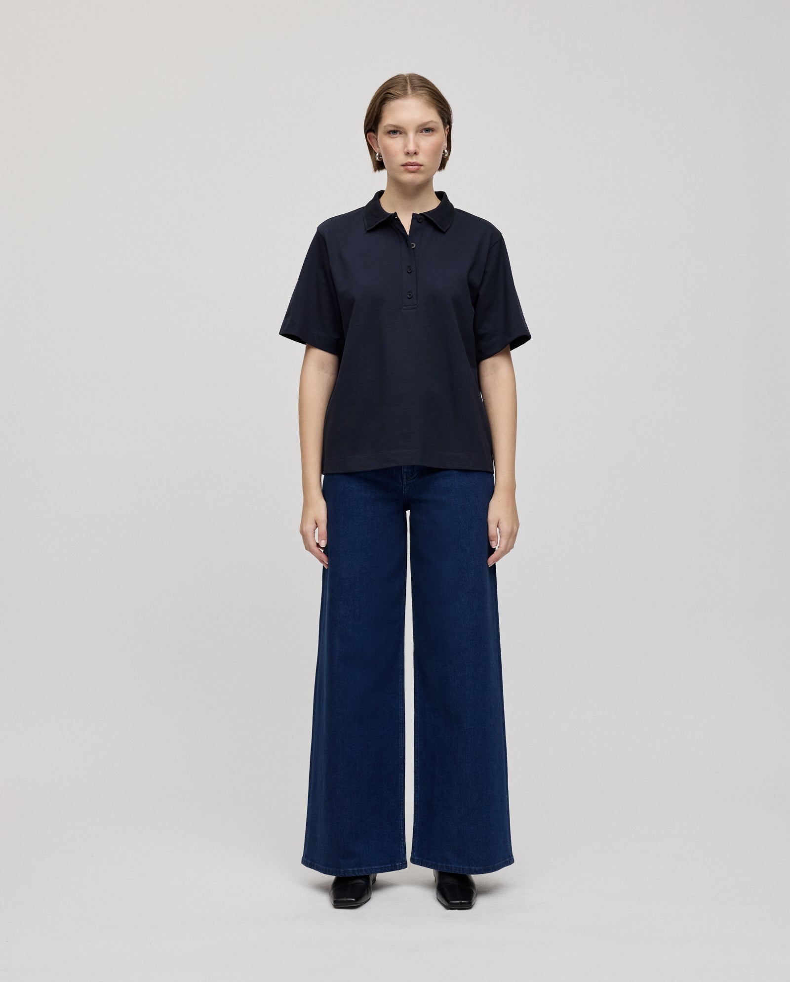 A woman stands facing forward, wearing the IVY OAK FABIOLA Top in black organic cotton and wide-leg blue jeans against a plain background.