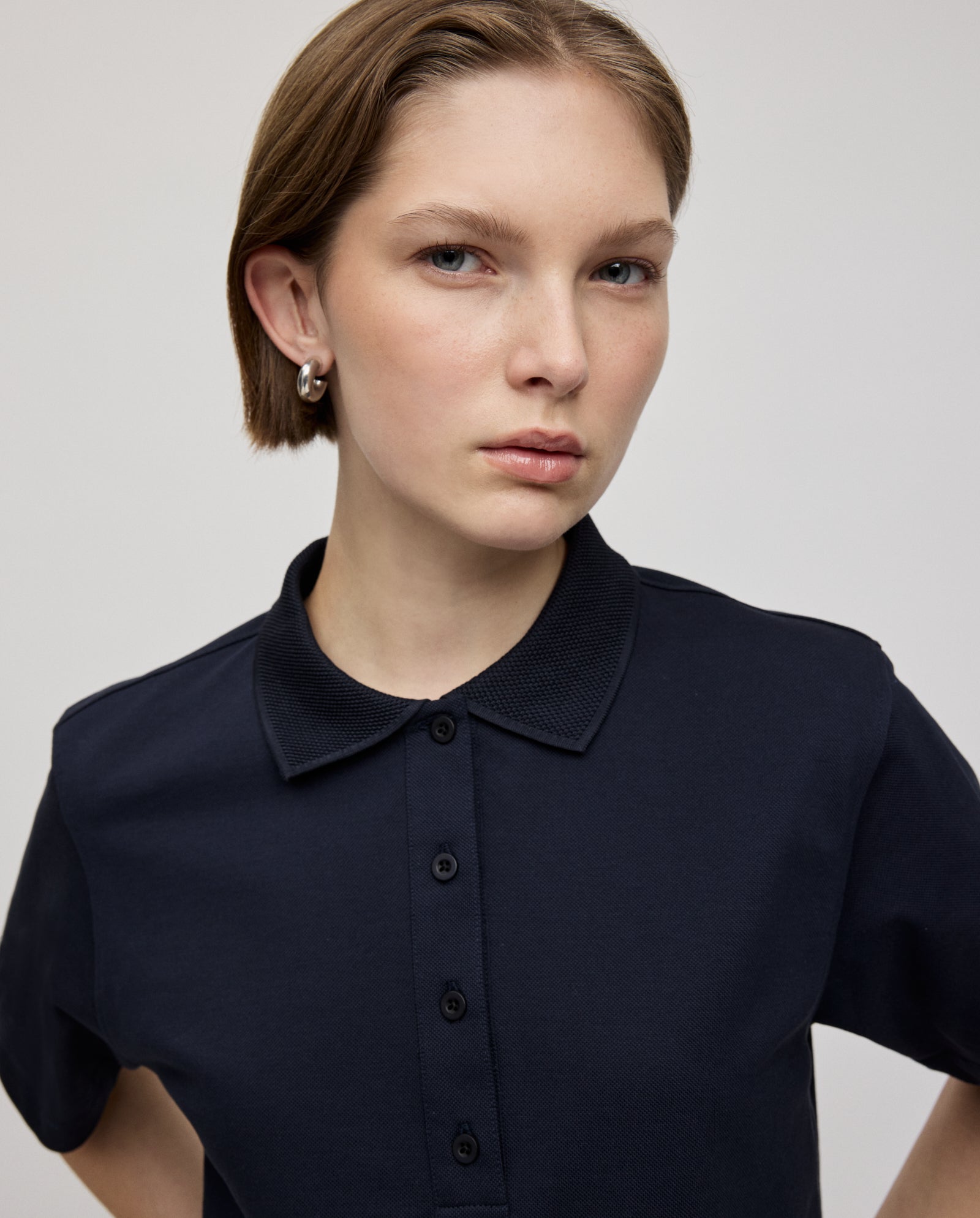 A young woman wearing the IVY OAK FABIOLA Top in navy organic cotton gazes ahead with a neutral expression against a plain background.