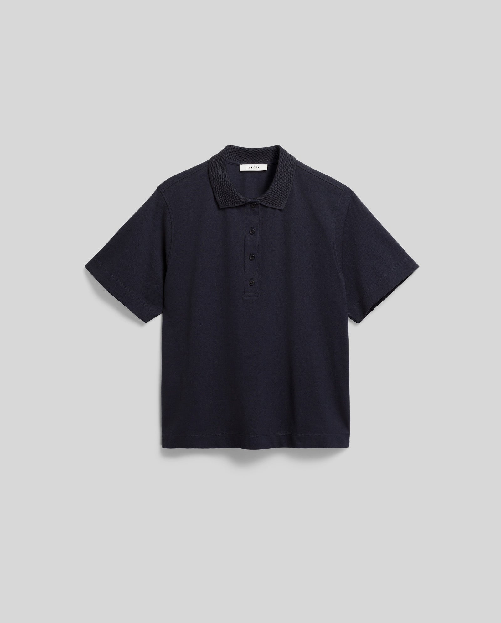 The FABIOLA Top by IVY OAK is a plain, short-sleeve, navy blue polo shirt crafted from organic cotton with a collar and three buttons, shown against a gray background.